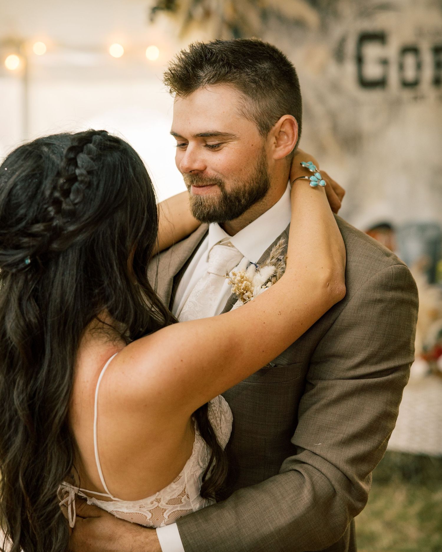 Smiling bearded groom in a tan suit and floral boutonniere embraces a bride with a long braided hairstyle during a wedding dance.