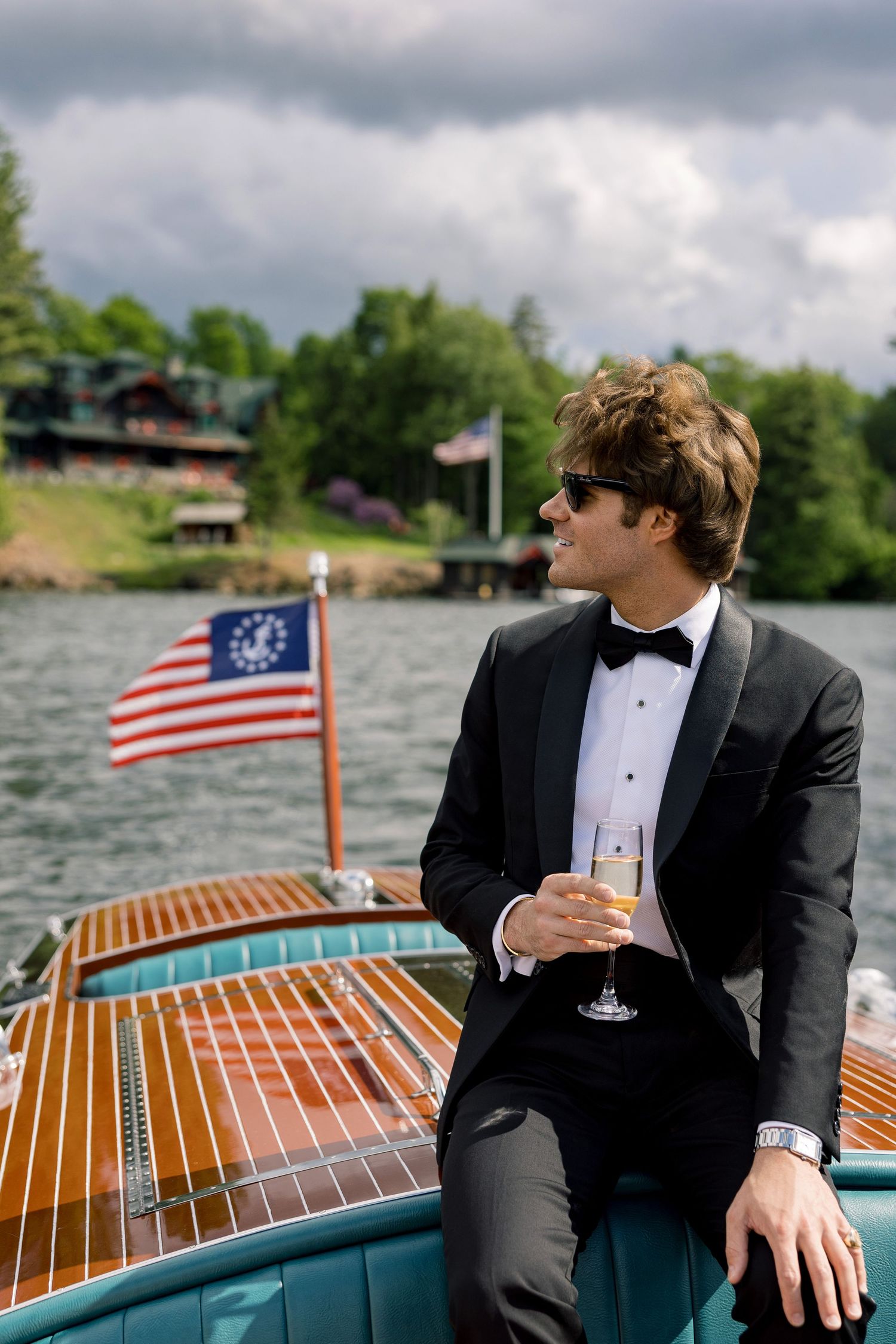Man in tuxedo holding champagne glass on wooden boat with American flag, lake and trees in background.
