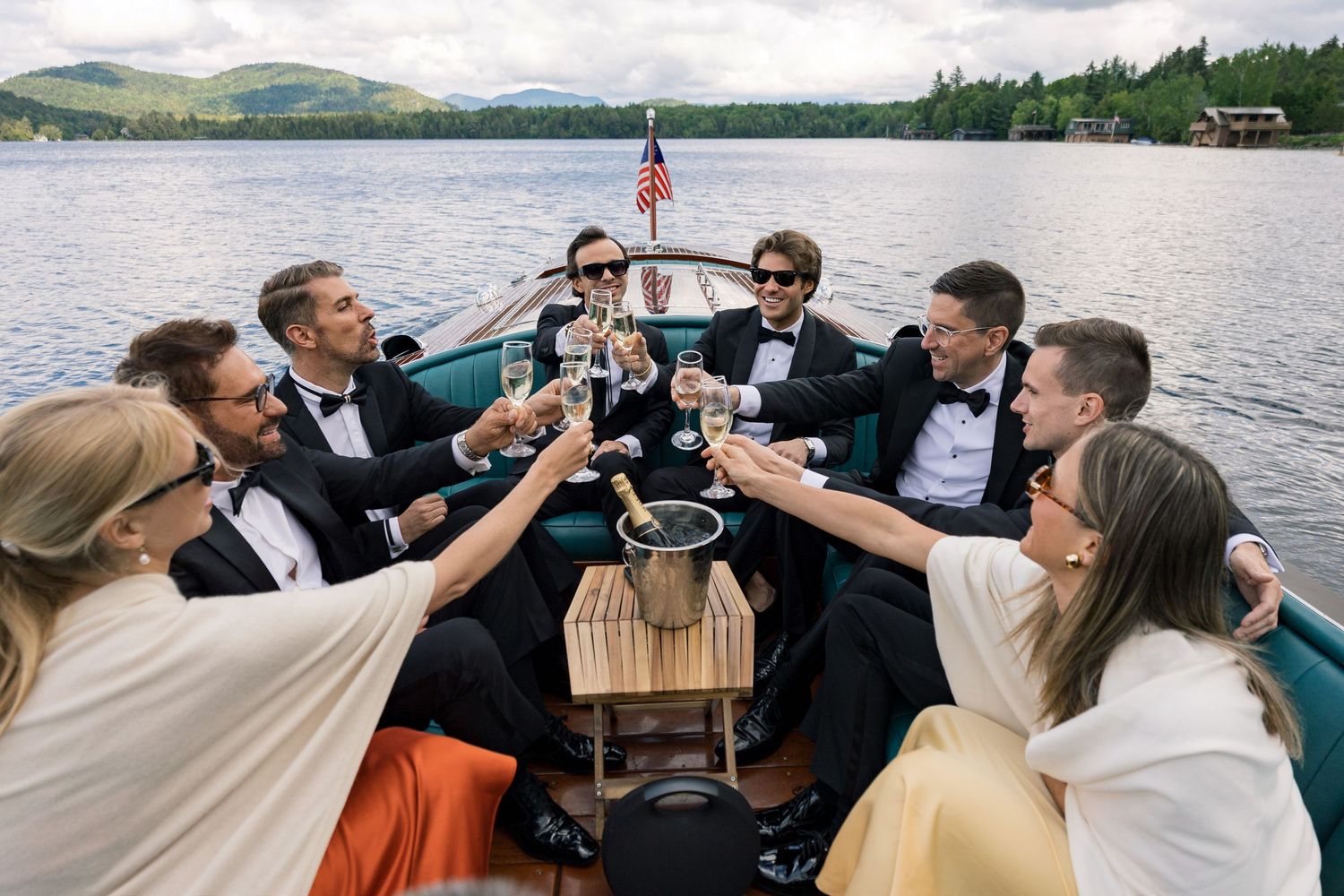 Elegant wedding party toasting champagne on a boat on a scenic lake, dressed in tuxedos and formal gowns.