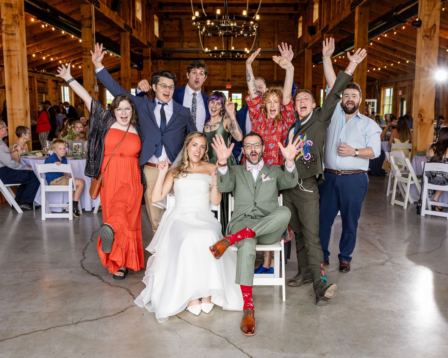 Joyful wedding party celebrating in a rustic barn venue, with bride and groom surrounded by cheering guests raising their arms.