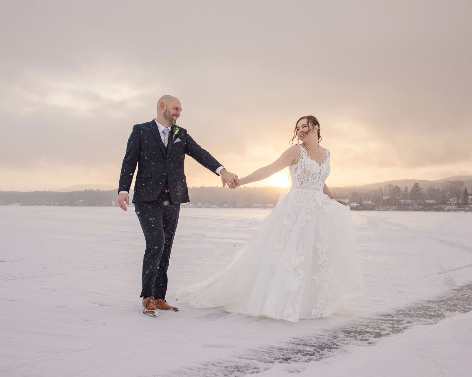 A couple in wedding attire holds hands while walking on a snowy landscape at sunset.