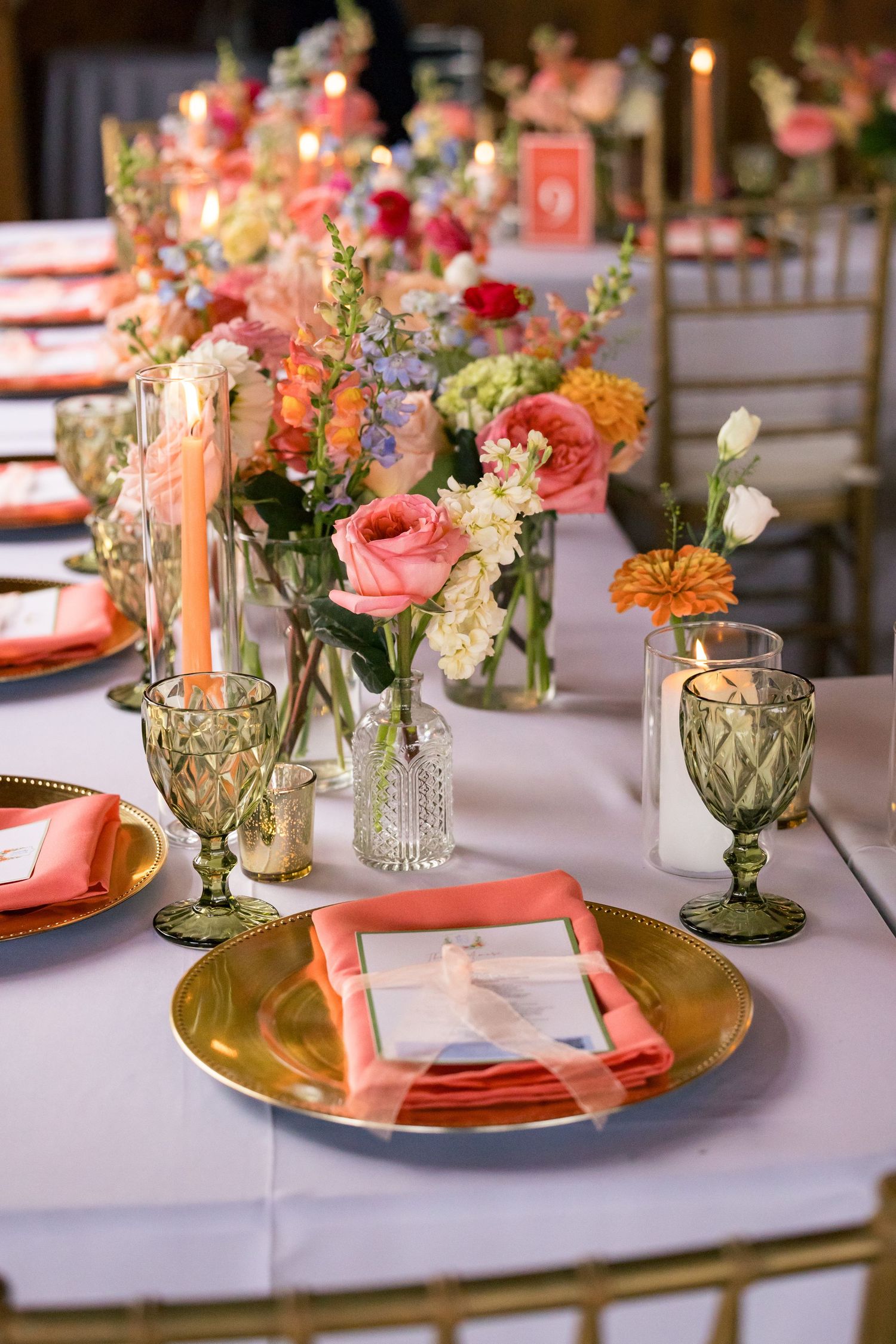 Elegant wedding reception table with gold chargers, coral napkins, green goblets, and colorful floral centerpieces.