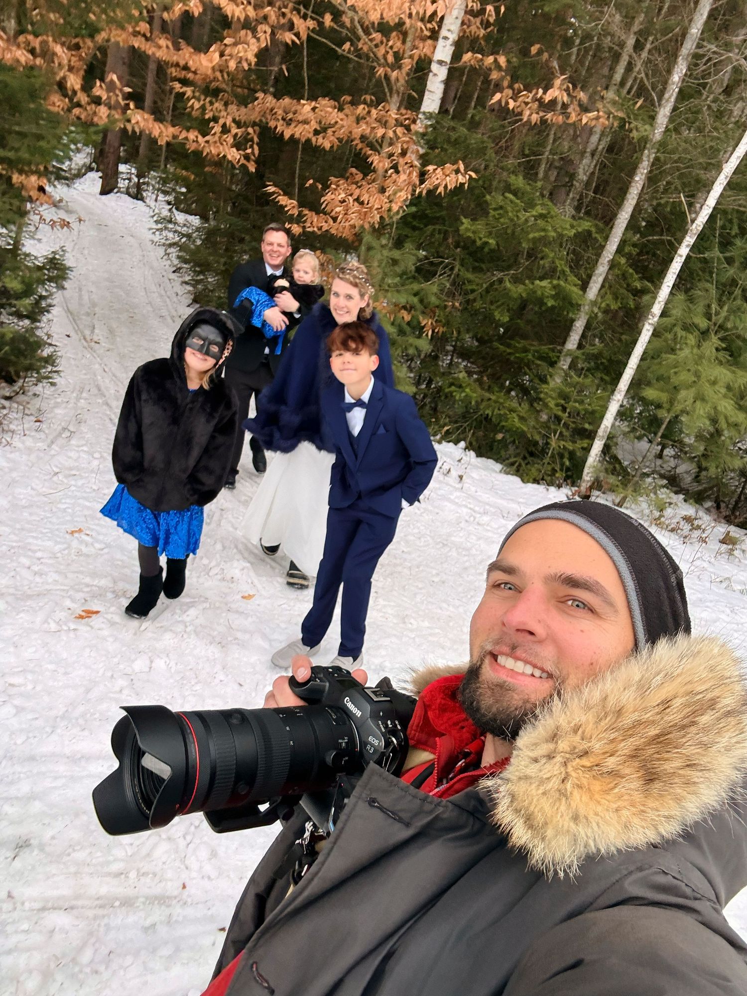 A photographer in winter gear takes a selfie with a professional camera on a snowy forest path.