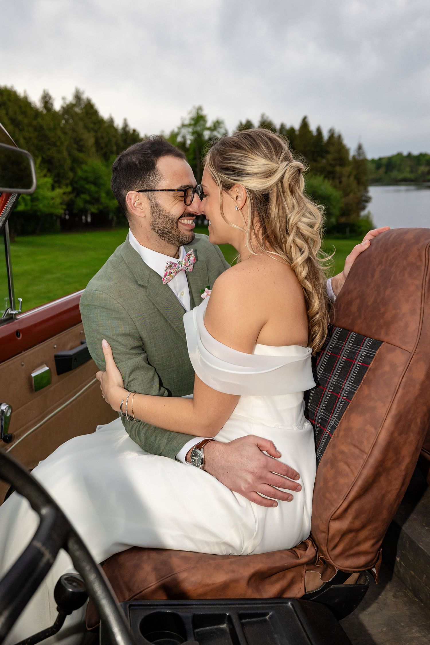 A bride and groom share a romantic moment sitting together in a vintage car with a scenic lake and trees in the background.