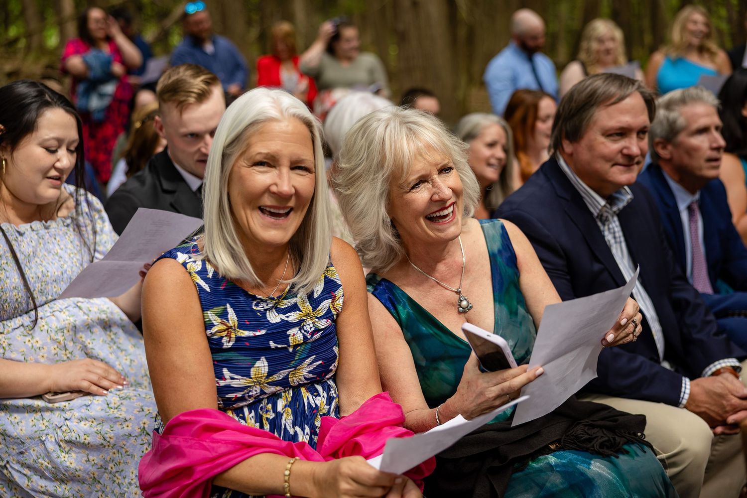 Two women laughing joyfully at an outdoor wedding ceremony, holding programs while seated among guests in formal attire.