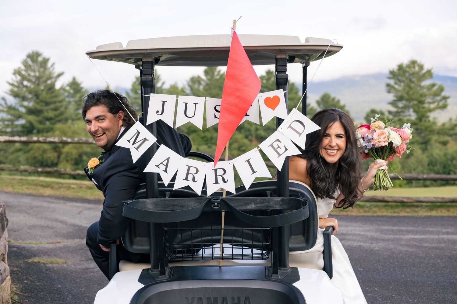 Newlyweds riding a Yamaha golf cart decorated with a Just Married banner and red flag at an outdoor wedding.