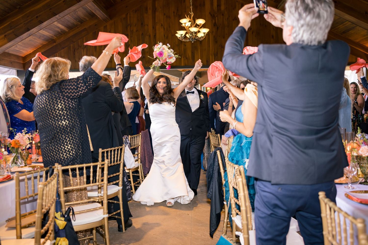 Bride and groom walk through cheering guests waving pink fans at elegant indoor wedding reception.