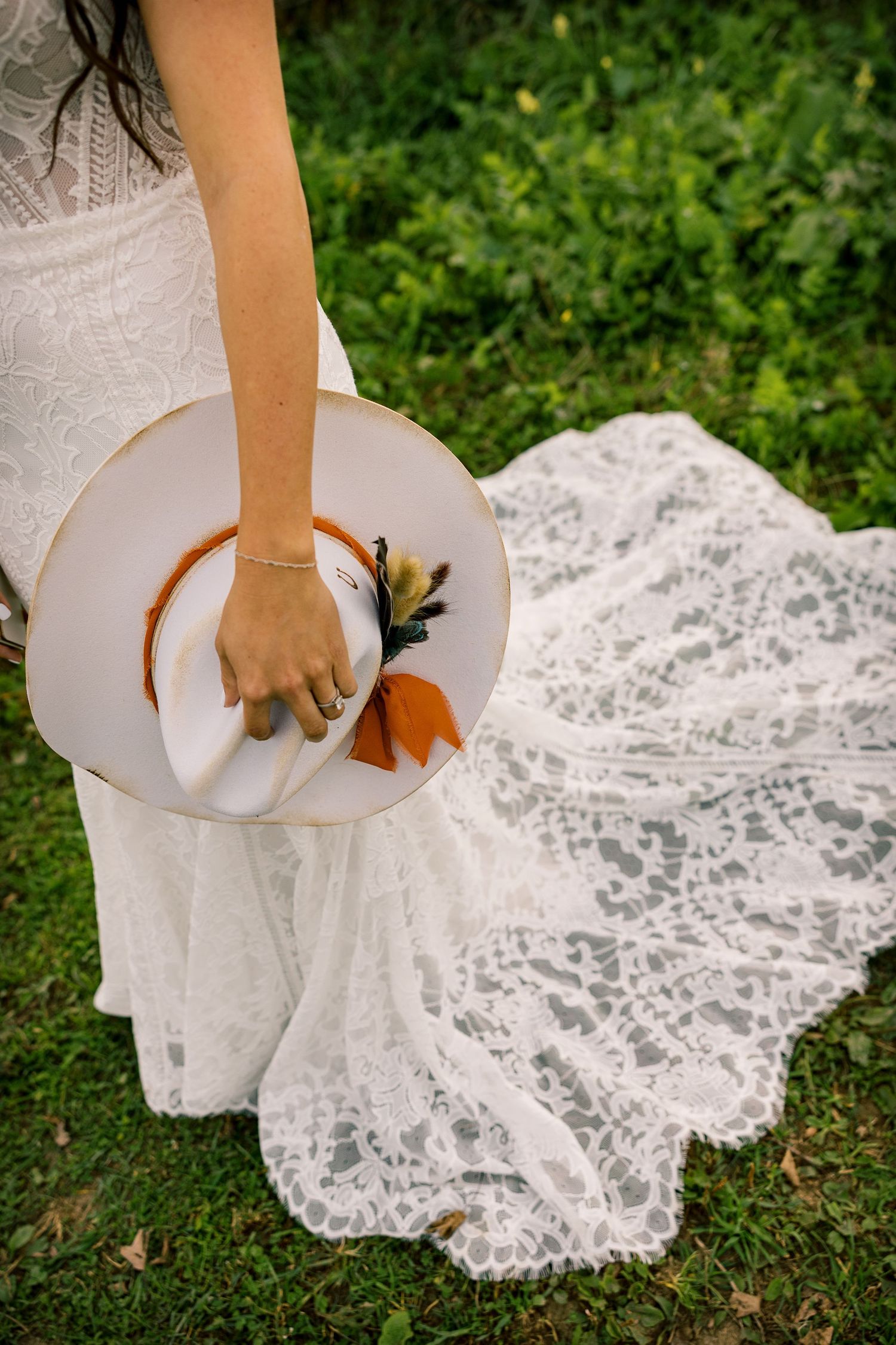 Bride in a flowing white lace wedding gown holds a cream boho hat with orange ribbon and feathers while standing on green grass.