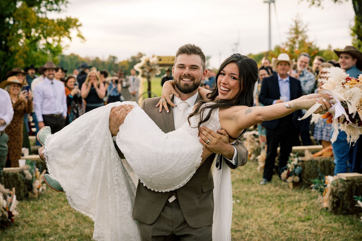 Groom in a gray suit carries his smiling bride in a white lace gown through an outdoor rustic wedding as guests in cowboy hats watch.