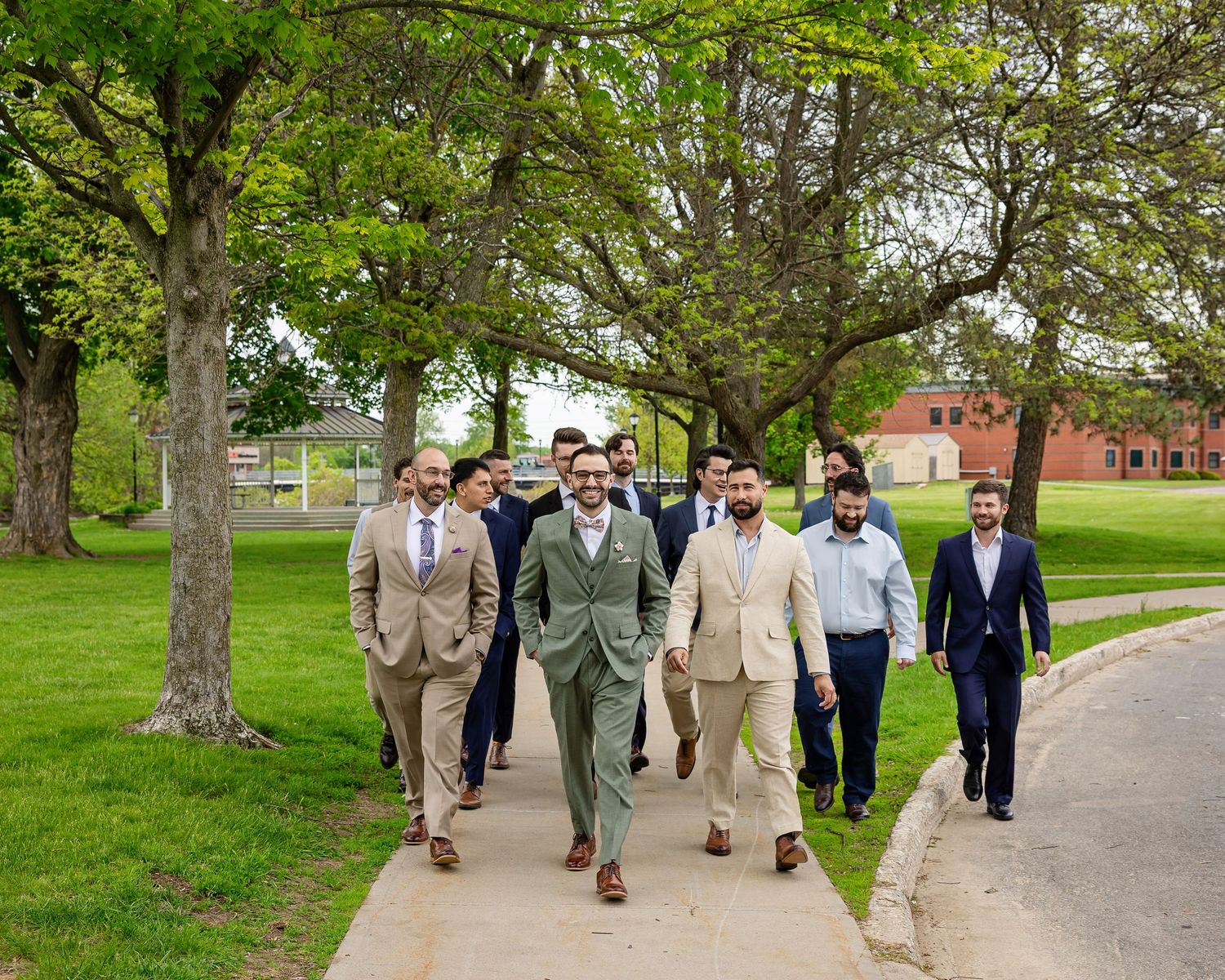 Group of well-dressed men in suits walking on a sidewalk through a green park on a sunny day.