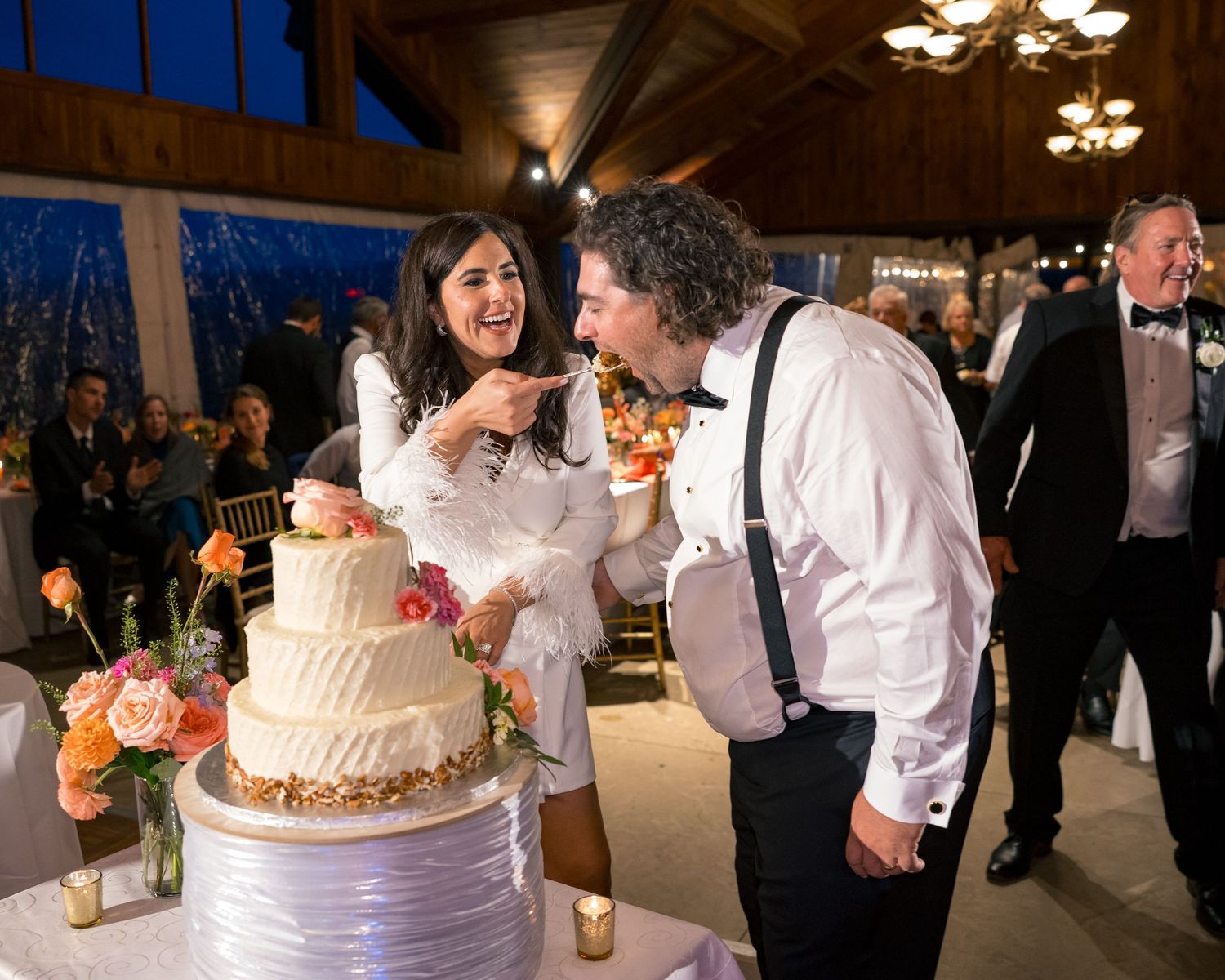 Bride and groom laughing while cutting their white wedding cake decorated with colorful flowers.