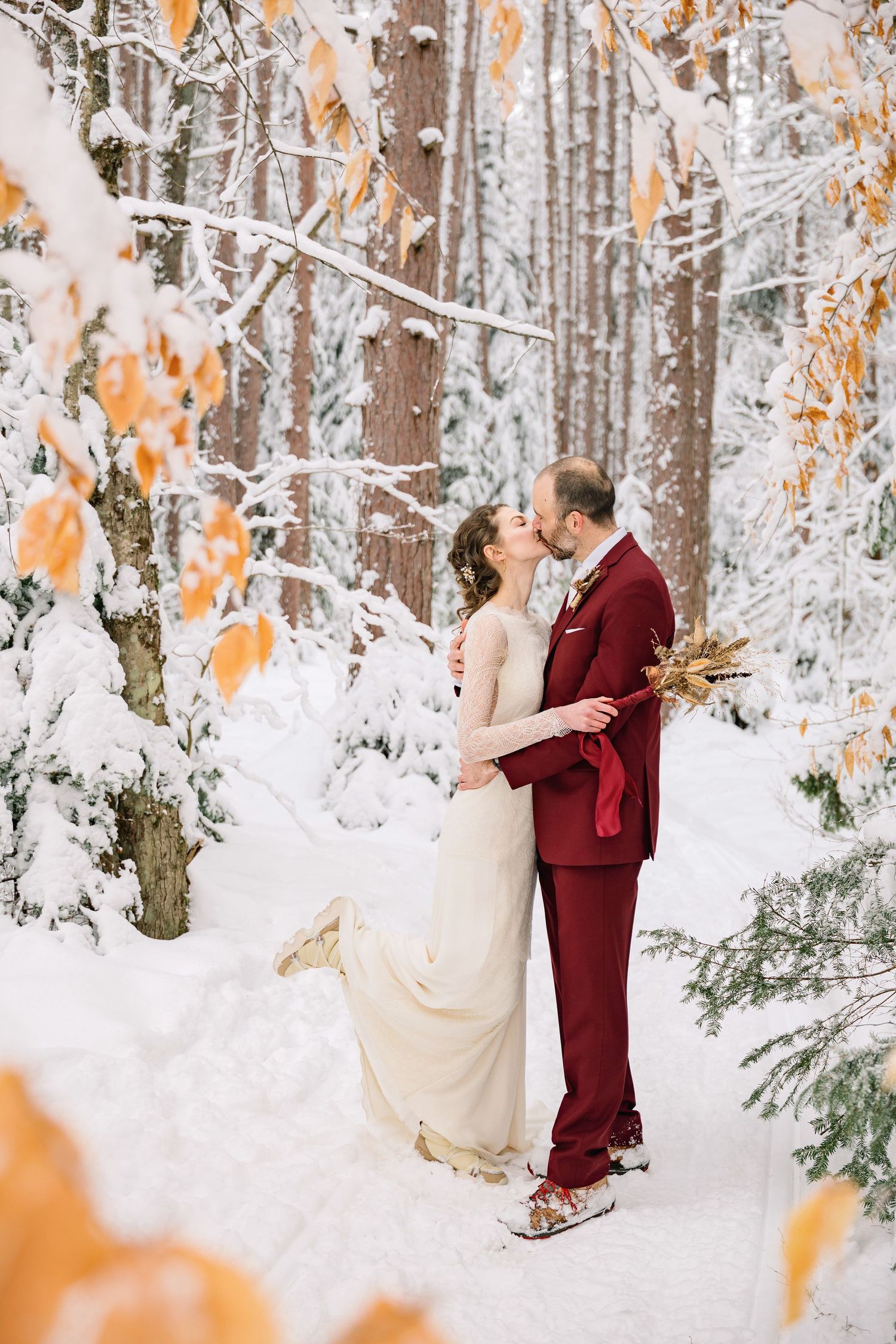 A couple shares a romantic moment in a snowy winter forest with orange autumn leaves still clinging to branches.