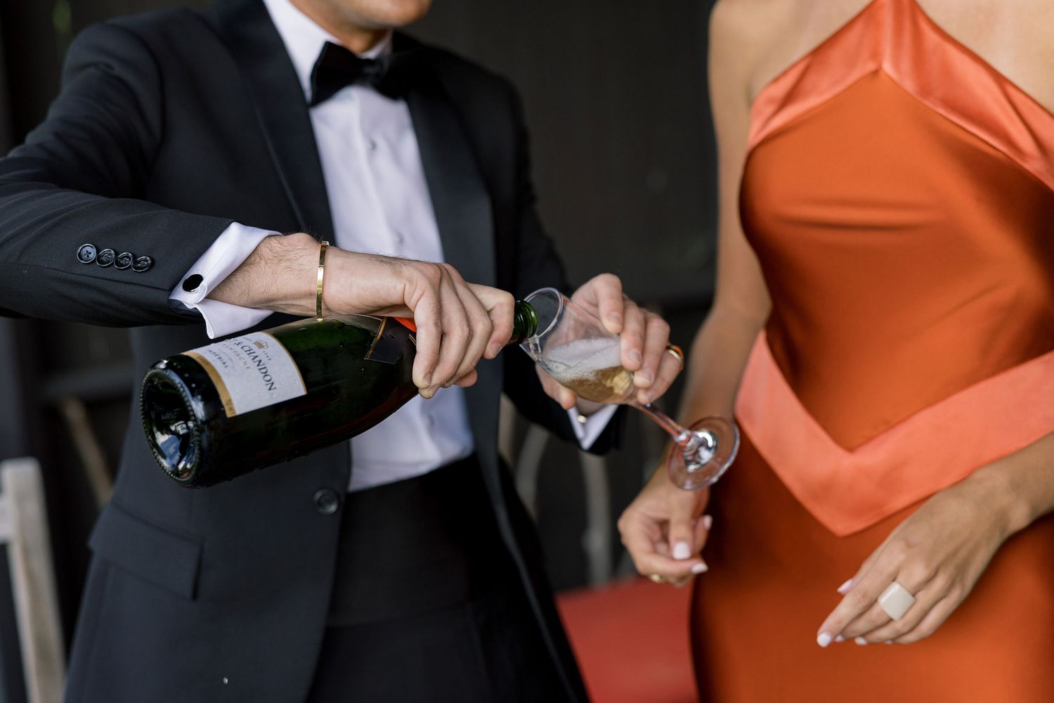 Man in tuxedo pouring champagne into a glass held by a woman in an elegant orange satin dress.