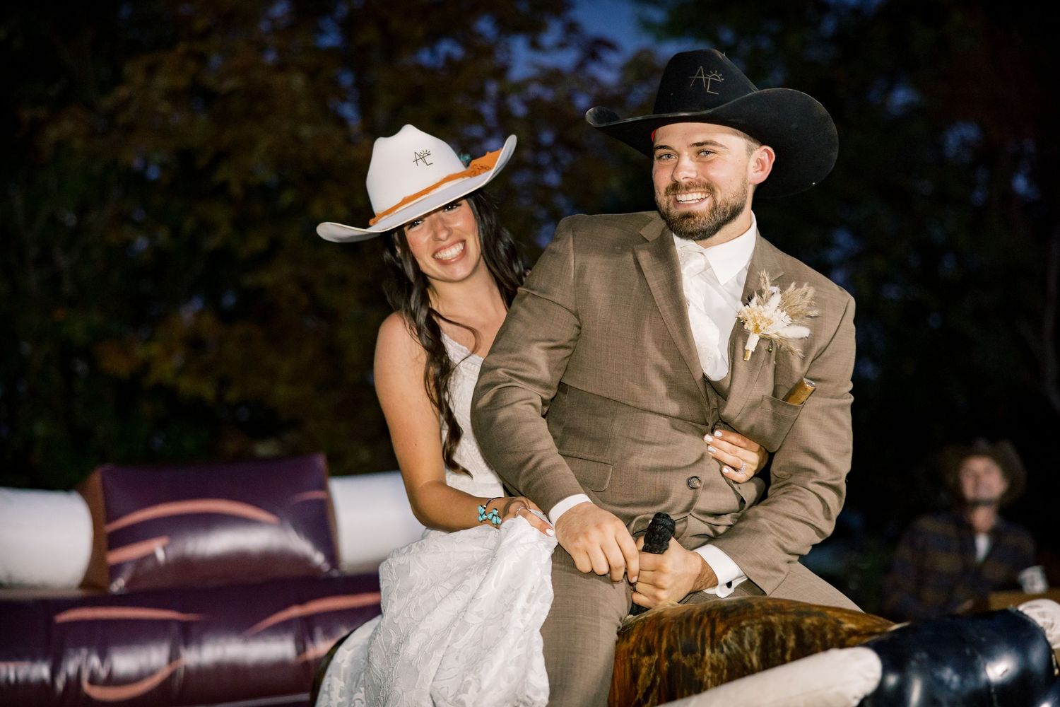 Bride in white dress and cowboy hat and groom in tan suit and black hat laugh together while riding a mechanical bull at their wedding.