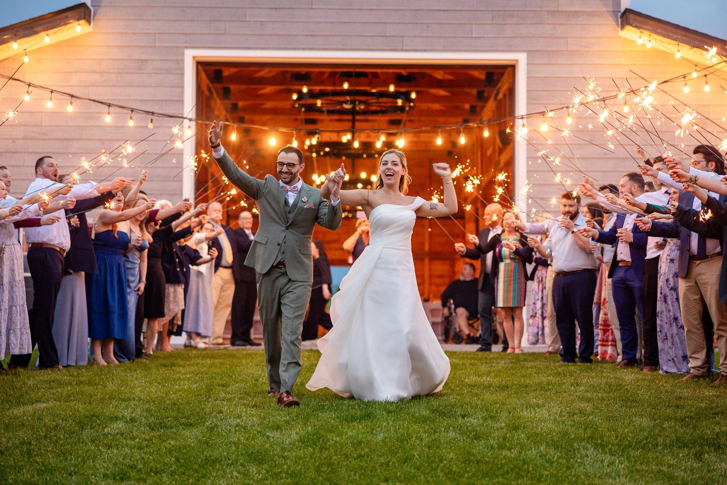 Bride and groom exit wedding reception through sparkler tunnel outside rustic barn venue at dusk.