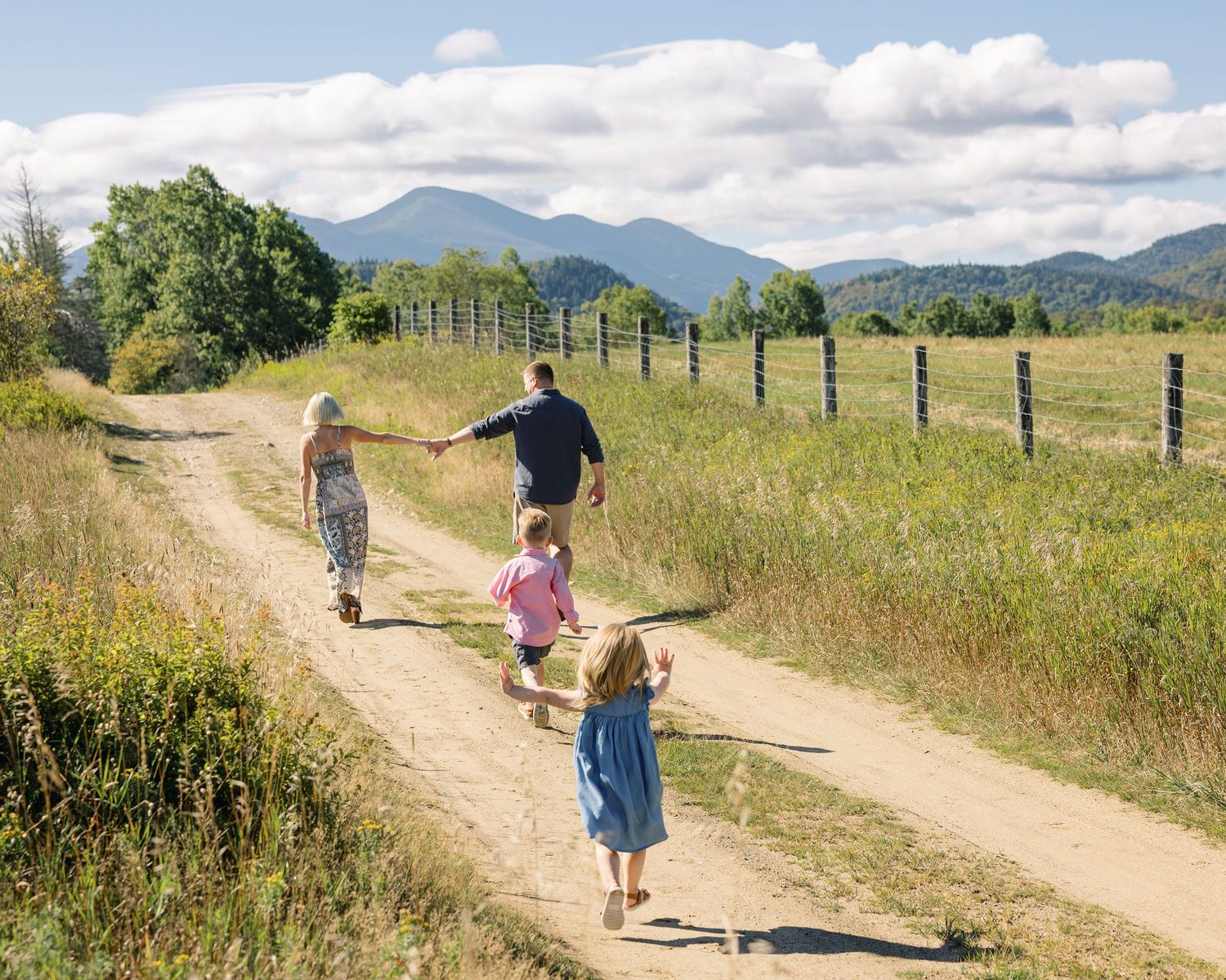 Hikers walk along a winding dirt path through grassy meadows with mountains visible in the distance on a sunny day.