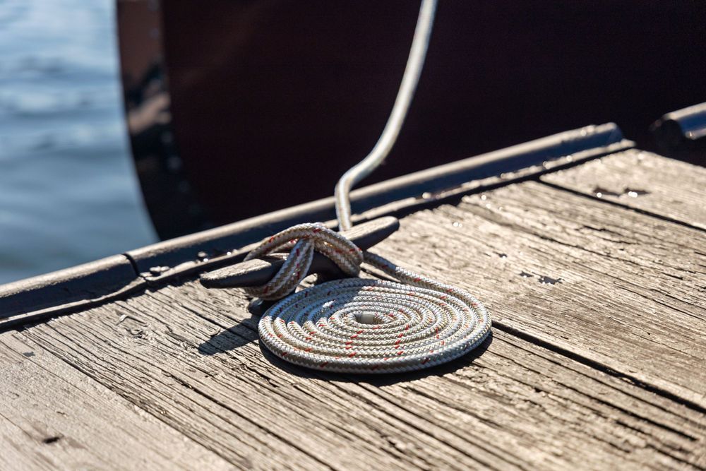A black boat cleat with rope tied around it on a weathered wooden dock near water.