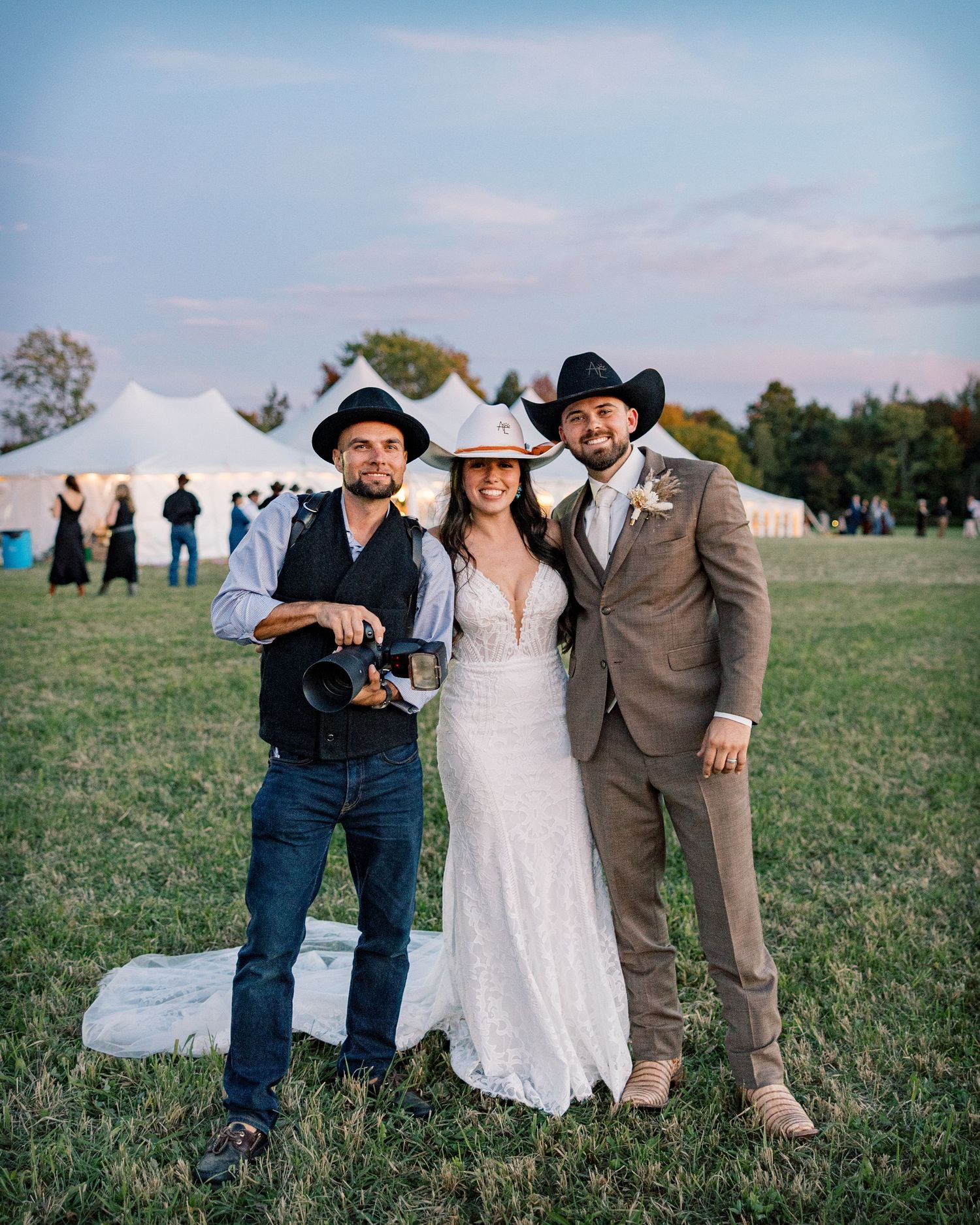 Bride in a lace wedding gown and groom in a tan suit, both in cowboy hats, pose with a photographer on a grassy field near white tents.