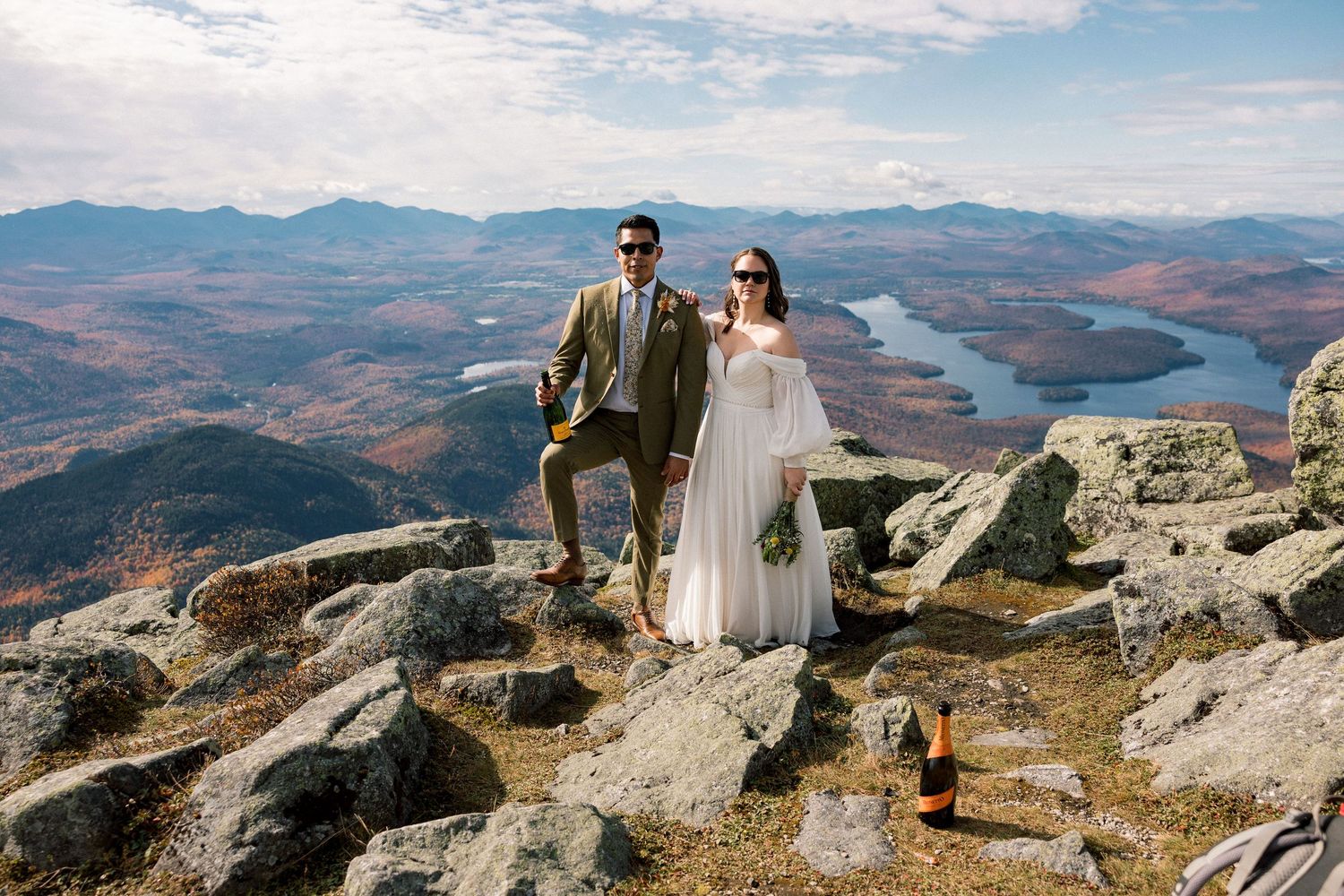 A couple stands on a mountain summit overlooking a vast autumn landscape with distant peaks and valleys below.
