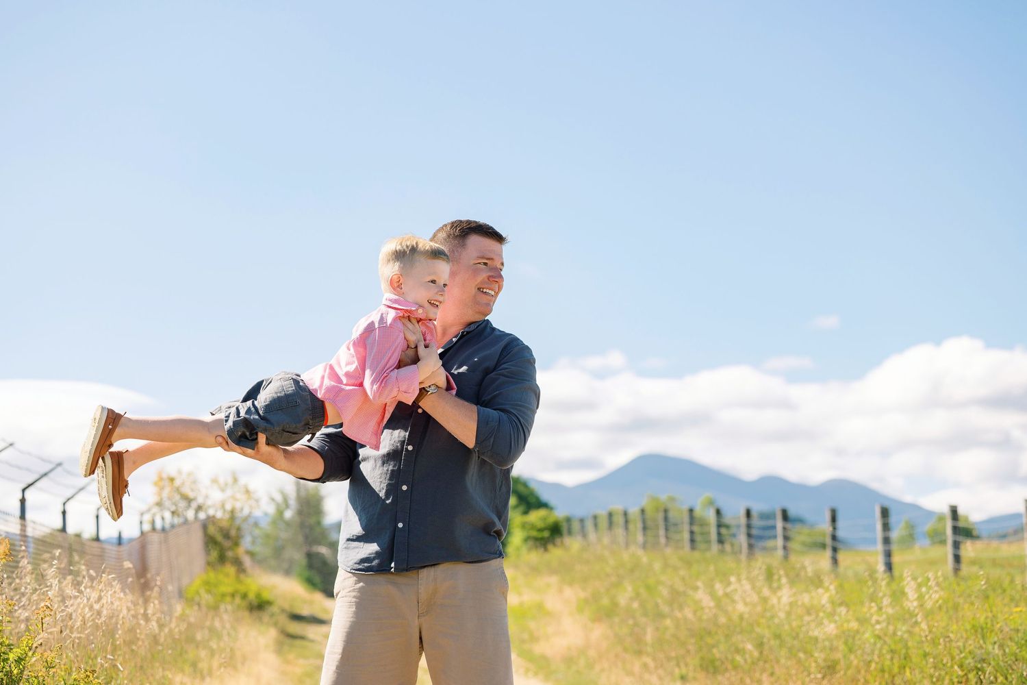 Two people share a playful moment in a sunny meadow with mountains in the background.