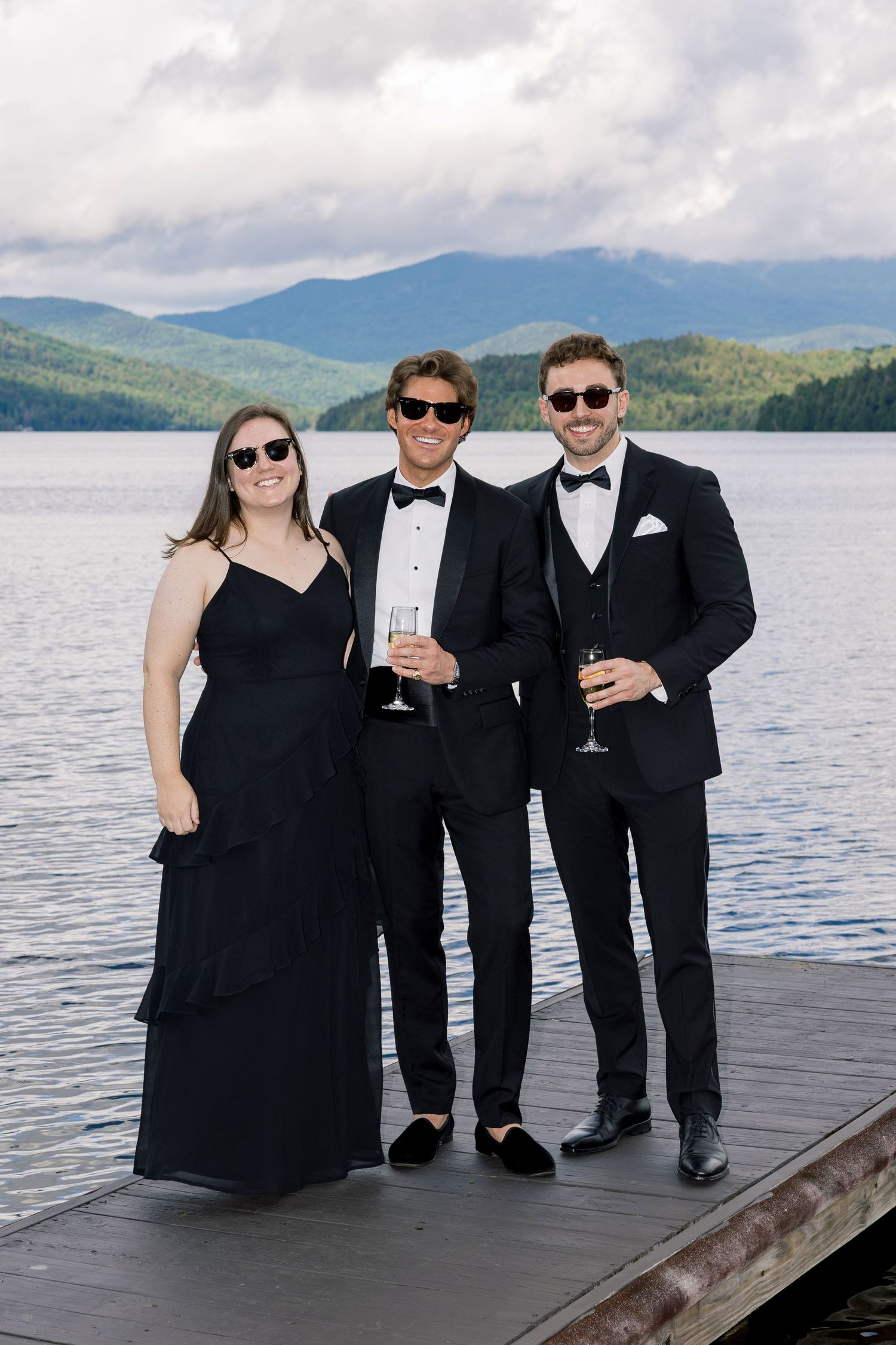 Three elegantly dressed guests in black formal wear pose on a lakeside dock with mountain views.