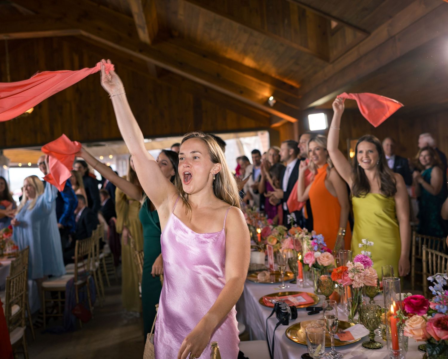 Guests wave red scarves in celebration at a colorful barn wedding reception with floral centerpieces.