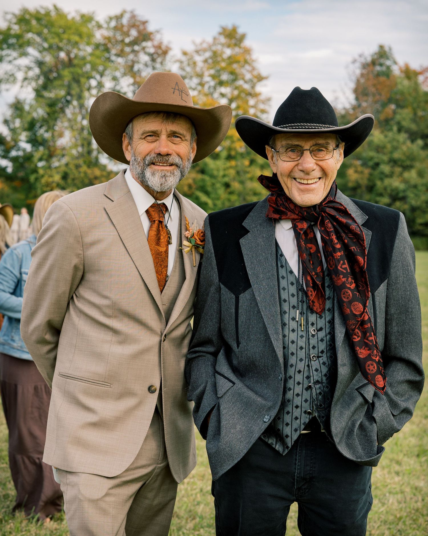 Two smiling men in cowboy hats and western-style attire pose together outdoors at a wedding, with autumn trees and guests behind them.