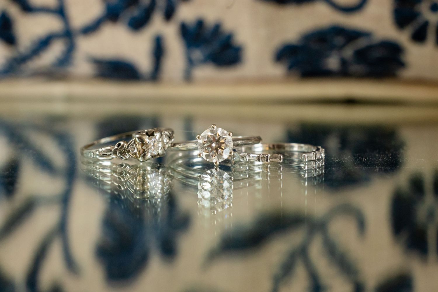Elegant diamond engagement rings and wedding bands reflected on a glass surface with blue floral background.
