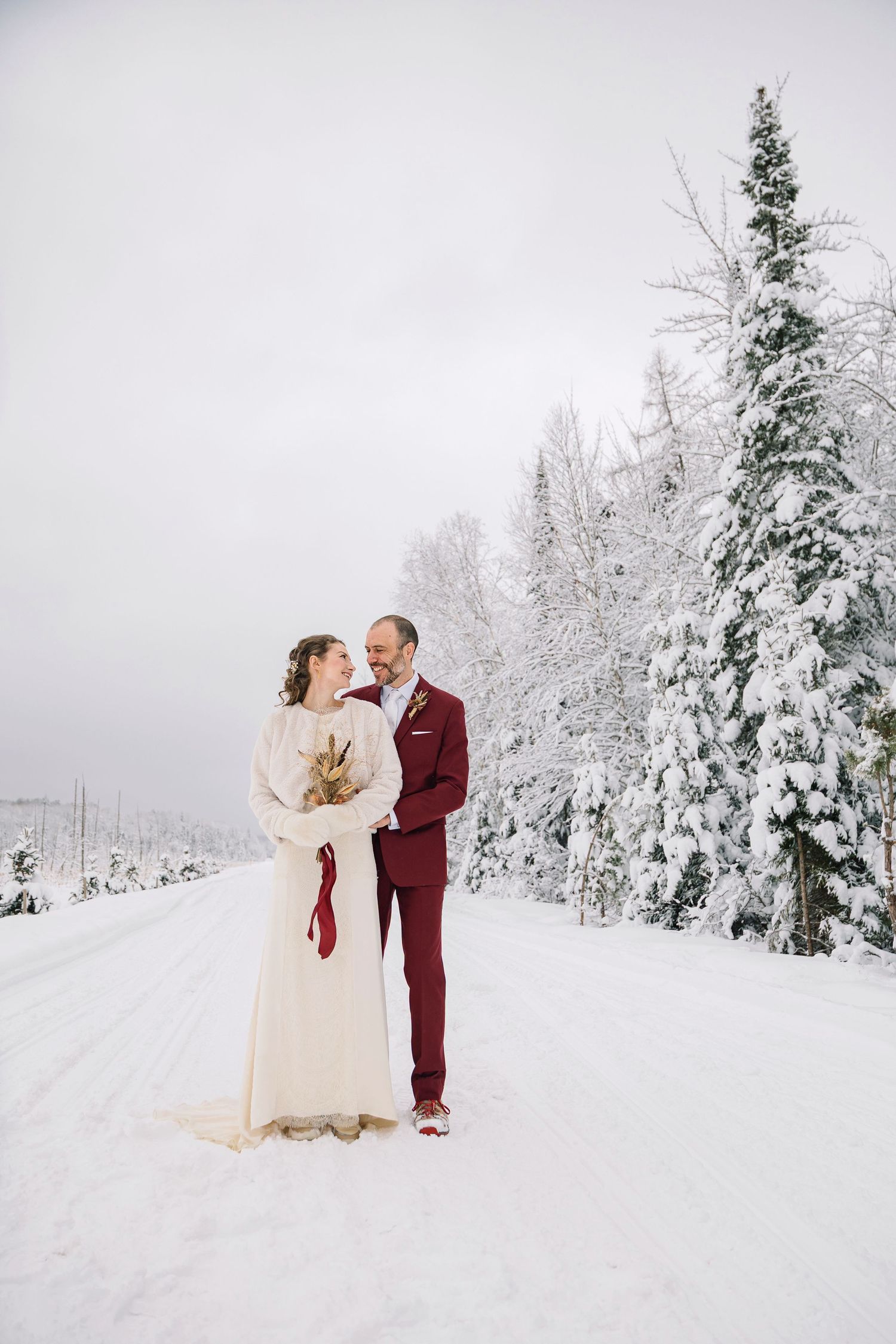 A couple in wedding attire embraces in a snowy winter wonderland with snow-covered evergreen trees in the background.