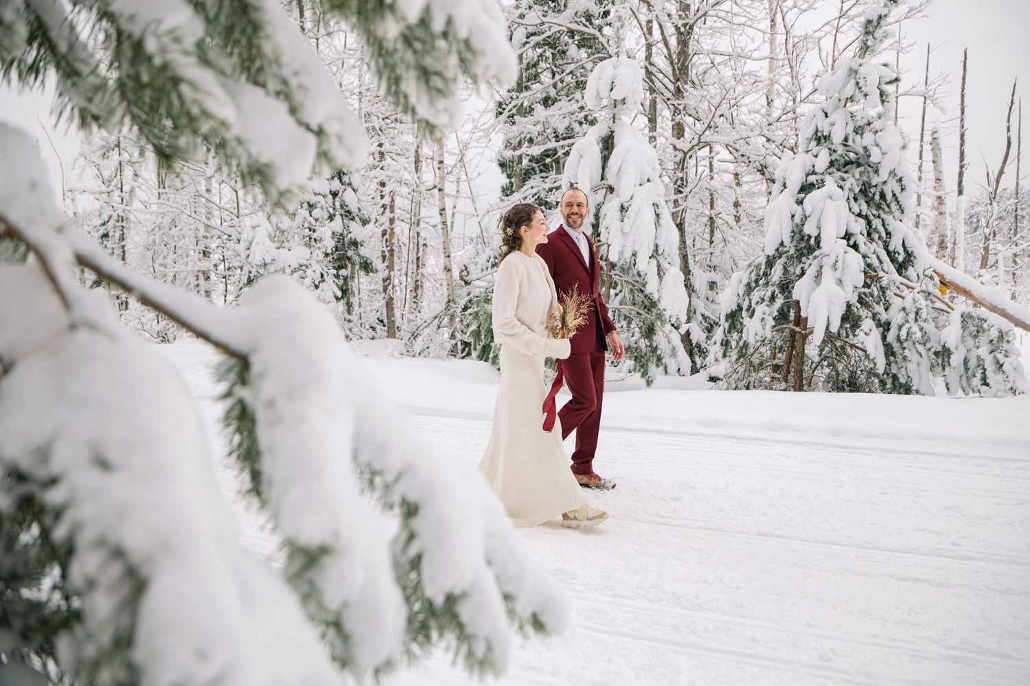 A couple in formal attire embraces in a snowy winter forest landscape with snow-covered evergreen trees.