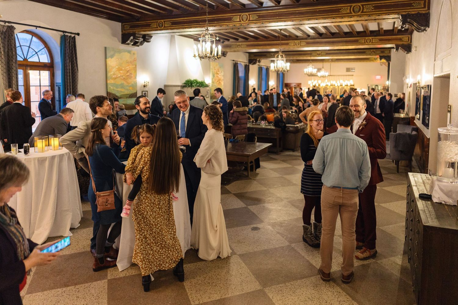 A crowded indoor event with people mingling in an elegant room featuring exposed wooden beams and a chandelier.