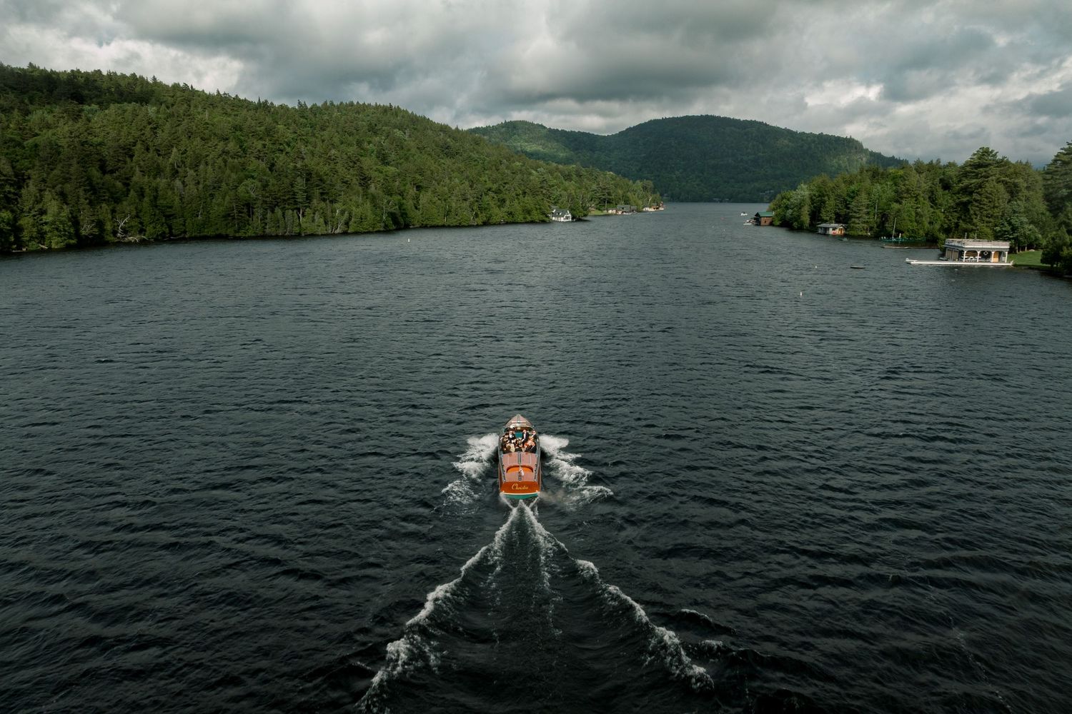Orange boat speeding down a calm mountain lake surrounded by lush green forested hills under cloudy skies.