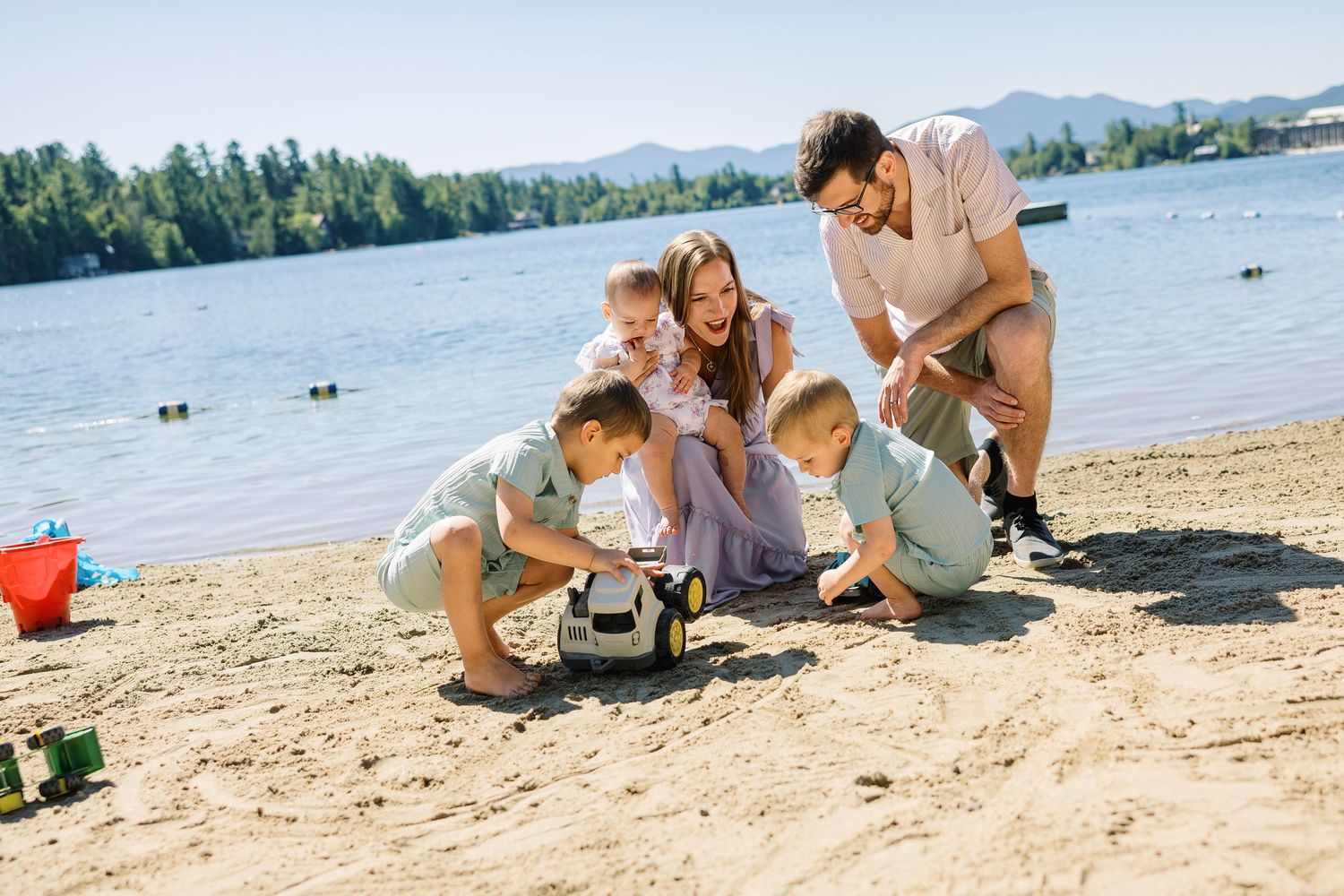 A family enjoys a sunny day together on a sandy beach by a lake surrounded by mountains and trees.