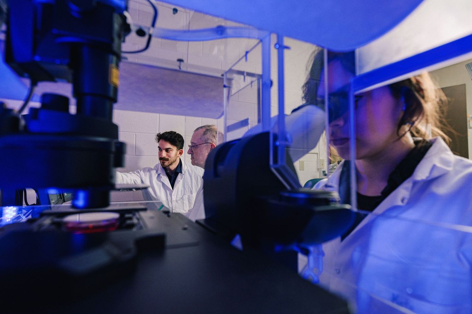 Medical professionals in protective gear work in a laboratory setting illuminated by blue lighting.