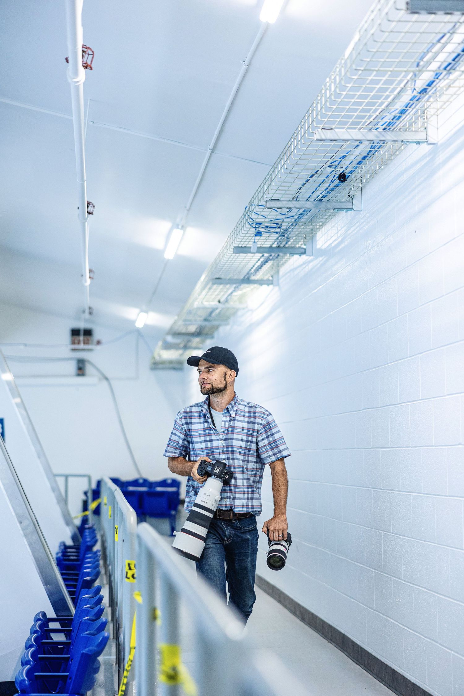 Person in plaid shirt and cap walking through bright industrial hallway with blue storage bins and white walls.