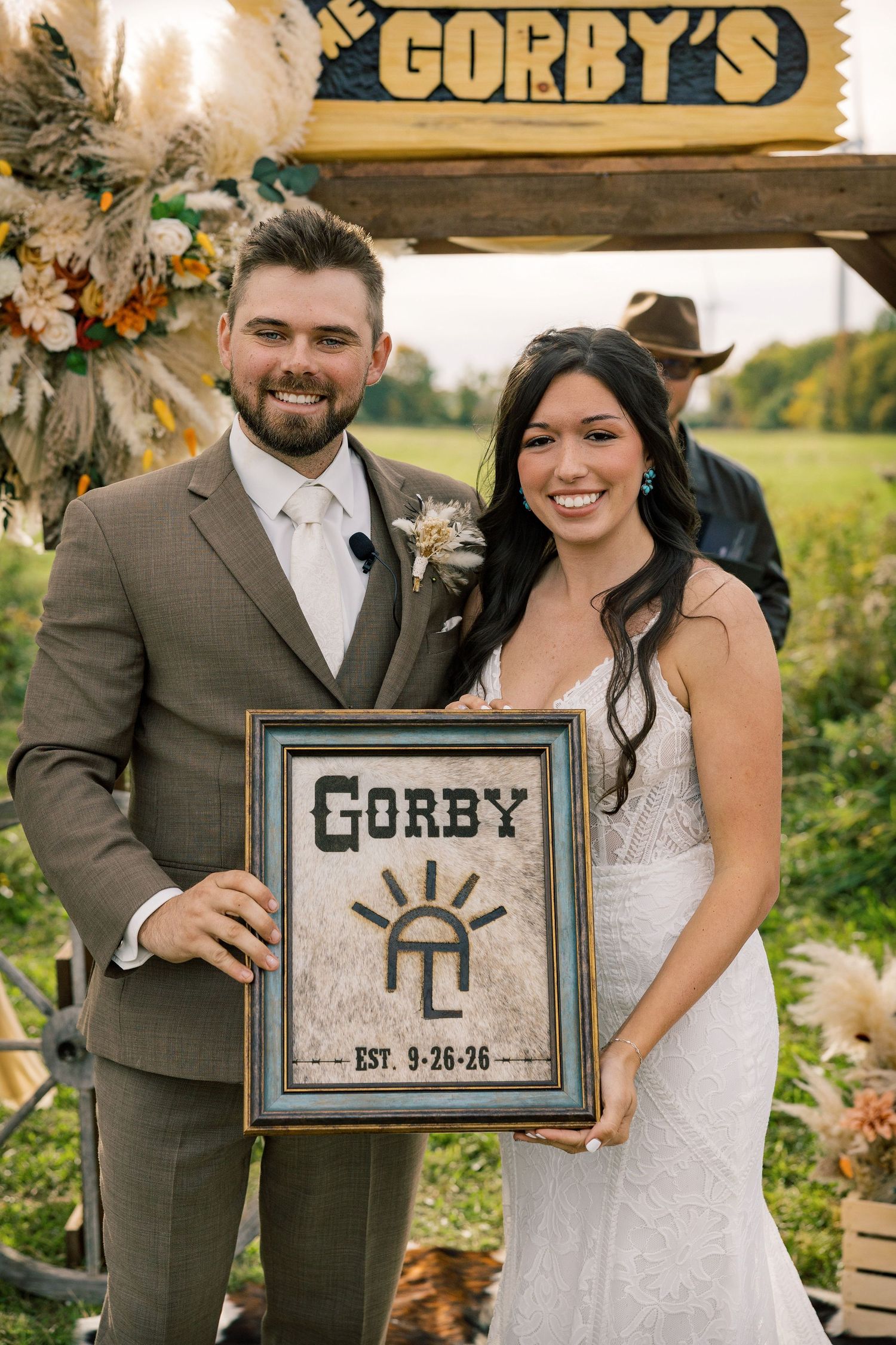 Smiling bride and groom hold a framed Gorby wedding sign dated 9-26-26 under a rustic wooden arch with floral decor.