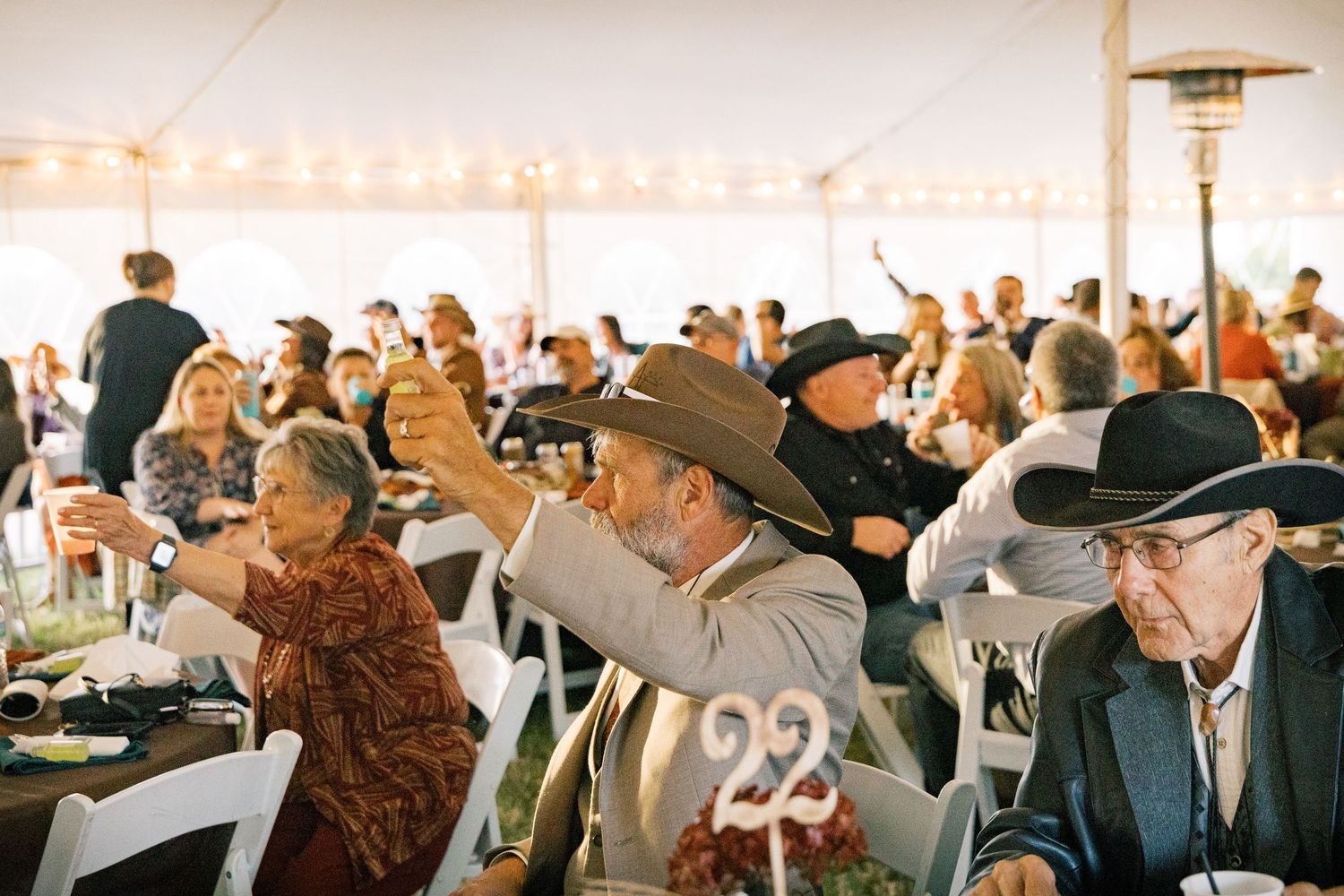 Guests in cowboy hats raise glasses in a toast at a festive banquet under a large white tent, with a number 22 sign on the table.