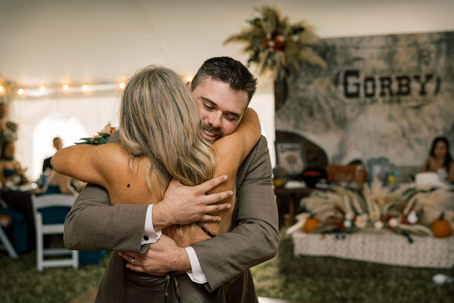 A groom in a gray suit hugs a woman with long blonde hair during a tented wedding reception with rustic decor and pumpkins.