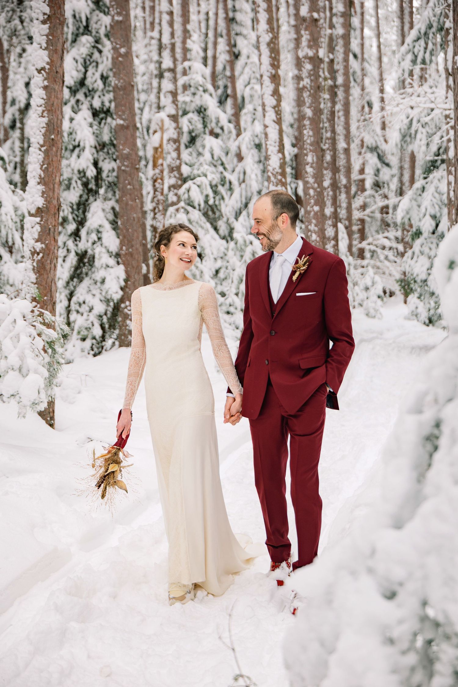 A couple in burgundy suit and cream dress share romantic moments in a snow-covered winter forest setting.
