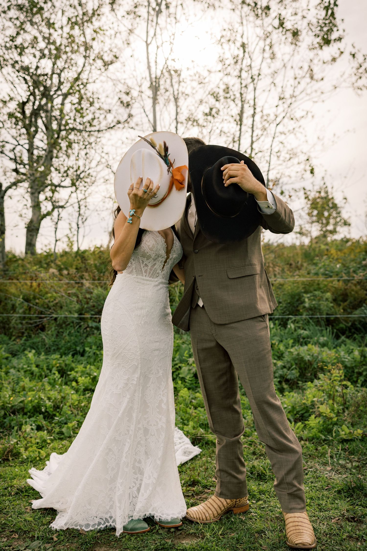 Bride in a lace wedding dress and groom in a tan suit hide their faces behind cowboy hats while kissing in a grassy field.