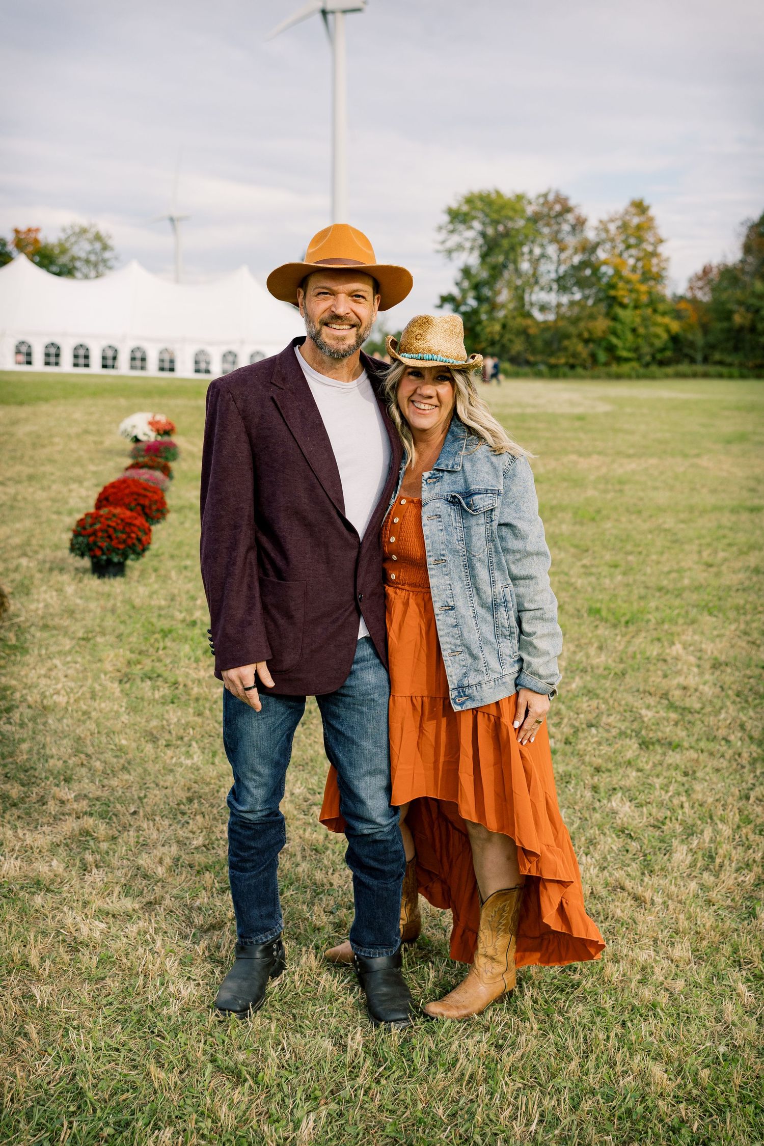 Smiling couple in cowboy hats and boots stand arm in arm on a grassy field near red mums, a white event tent and wind turbines.