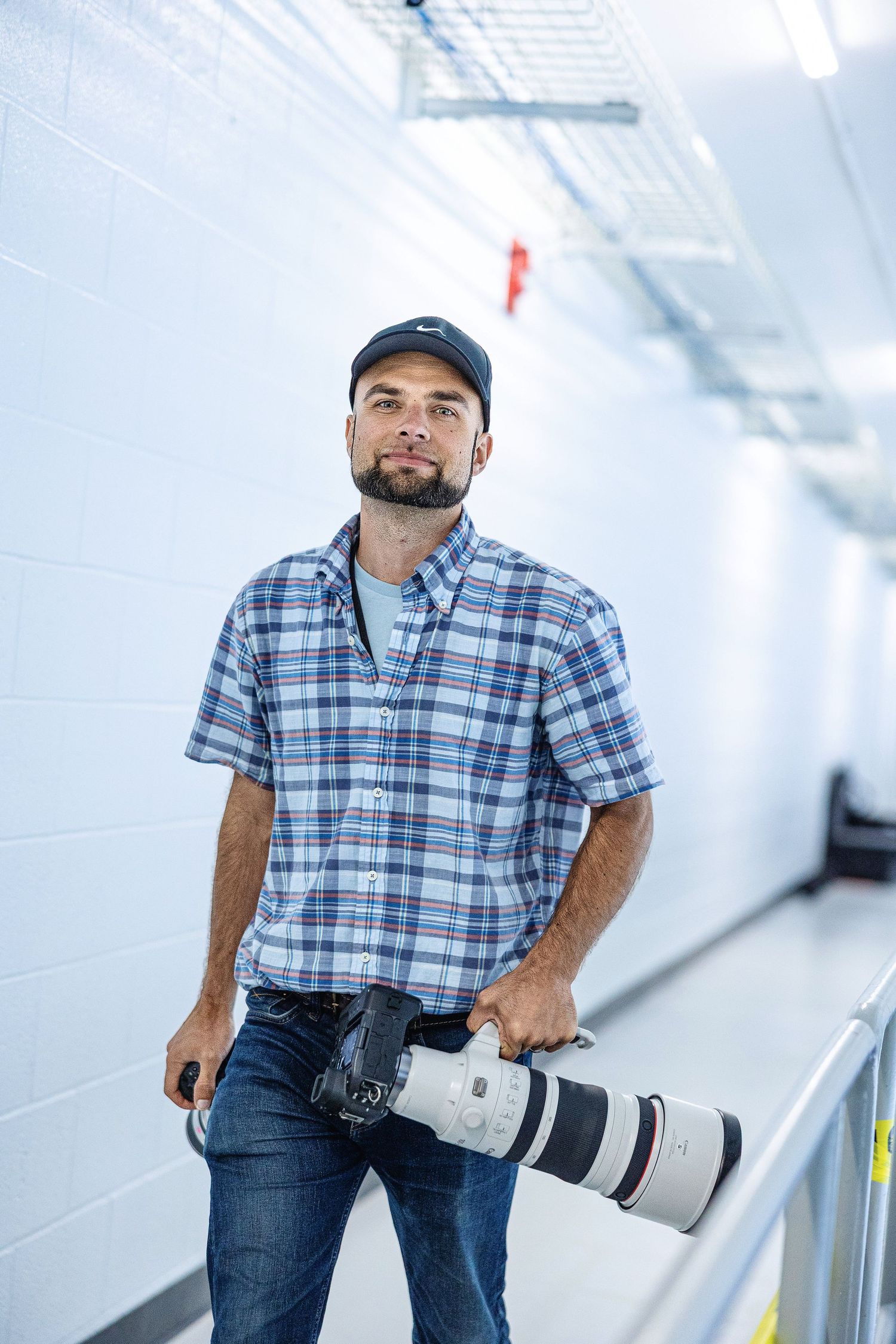 Professional photographer in blue plaid shirt holding telephoto lens in bright white hallway setting.