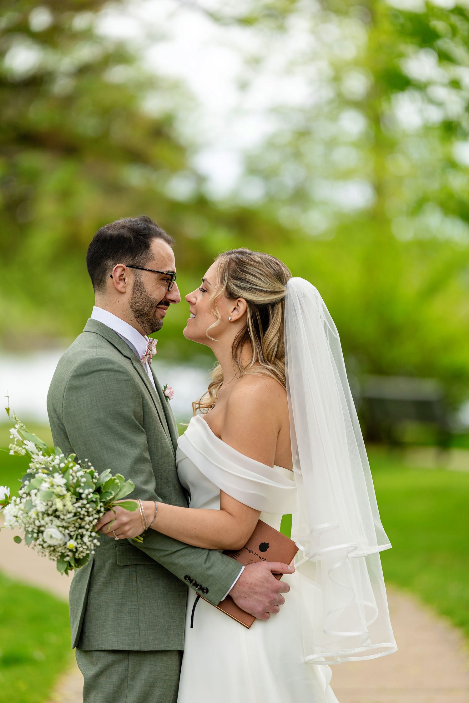A bride and groom share a romantic moment outdoors, with the groom in a sage suit and bride in a white off-shoulder gown with veil.