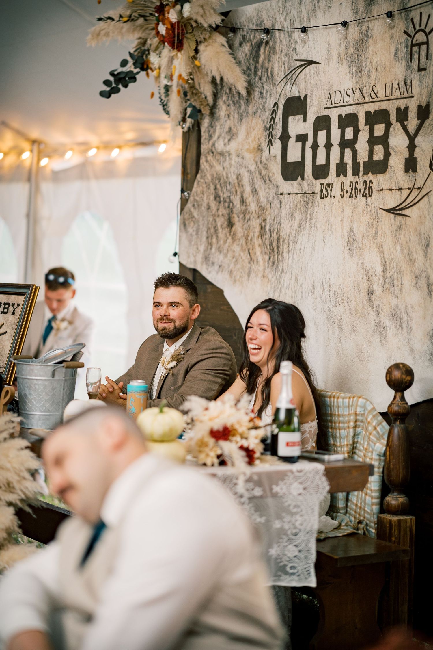Smiling couple sits at a rustic wedding head table with floral centerpieces and a Gorby family name banner behind them.
