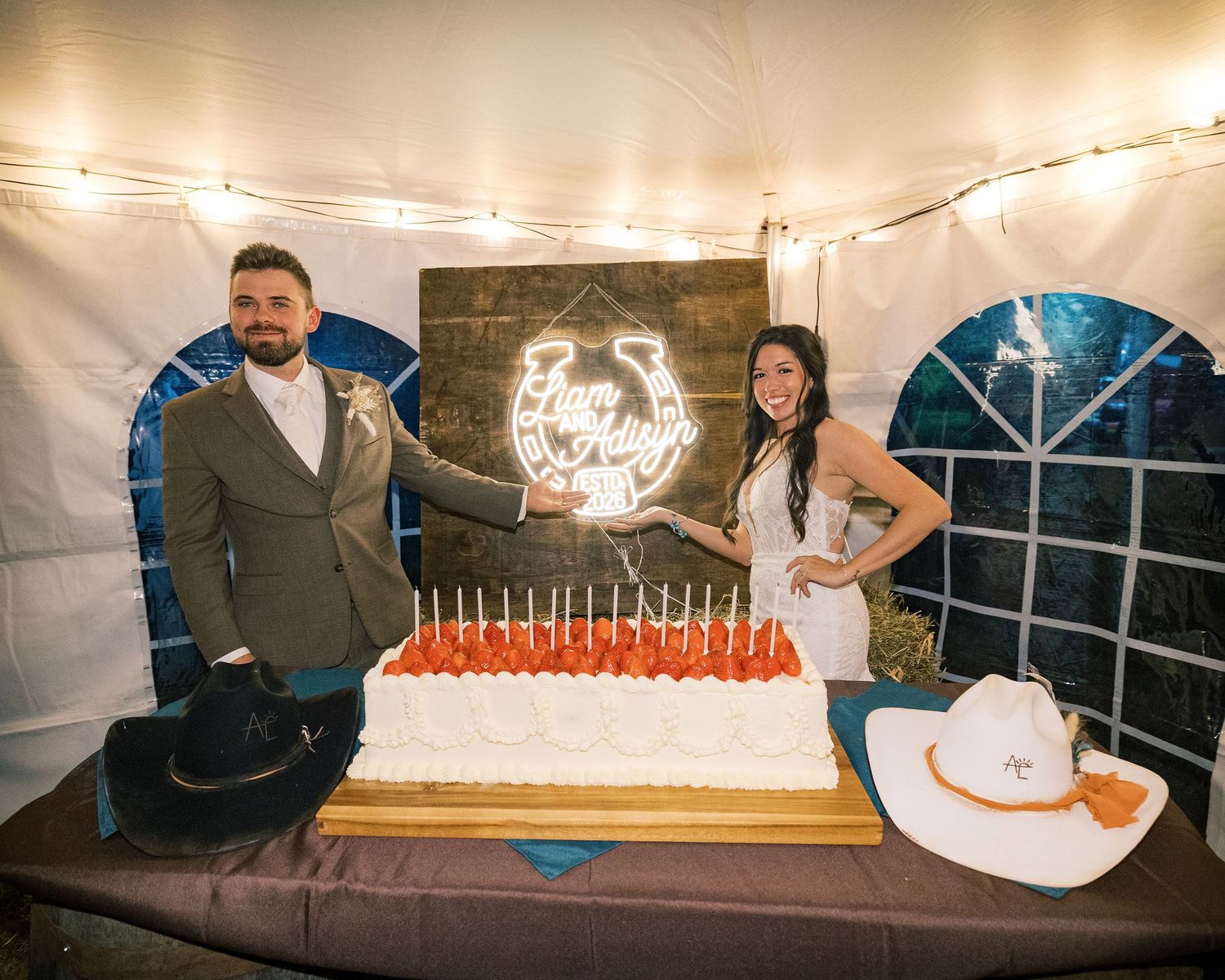 Smiling couple poses behind a large white wedding cake topped with red candles beside a neon horseshoe sign, with cowboy hats on the table.