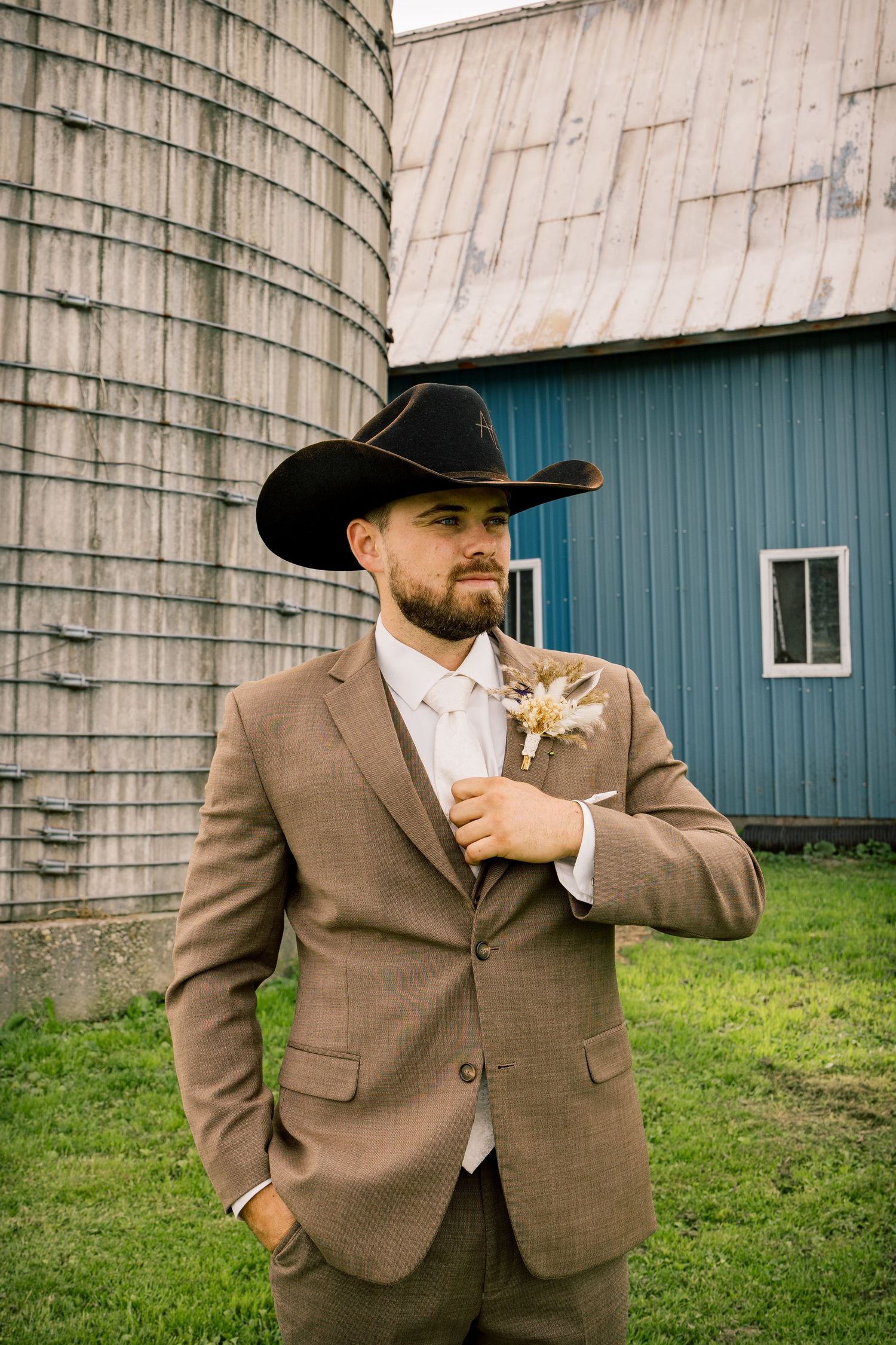 Groom in a tan suit and black cowboy hat stands on grass beside a weathered silo and blue barn, wearing a floral boutonniere.