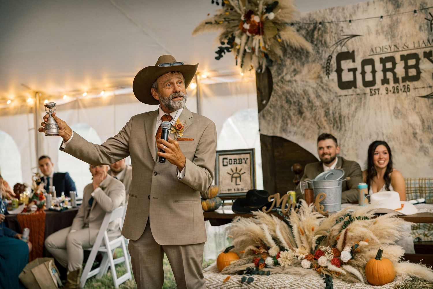 Man in a tan suit and cowboy hat gives a toast at a rustic tent wedding reception with pumpkins and wheat decor as guests watch.