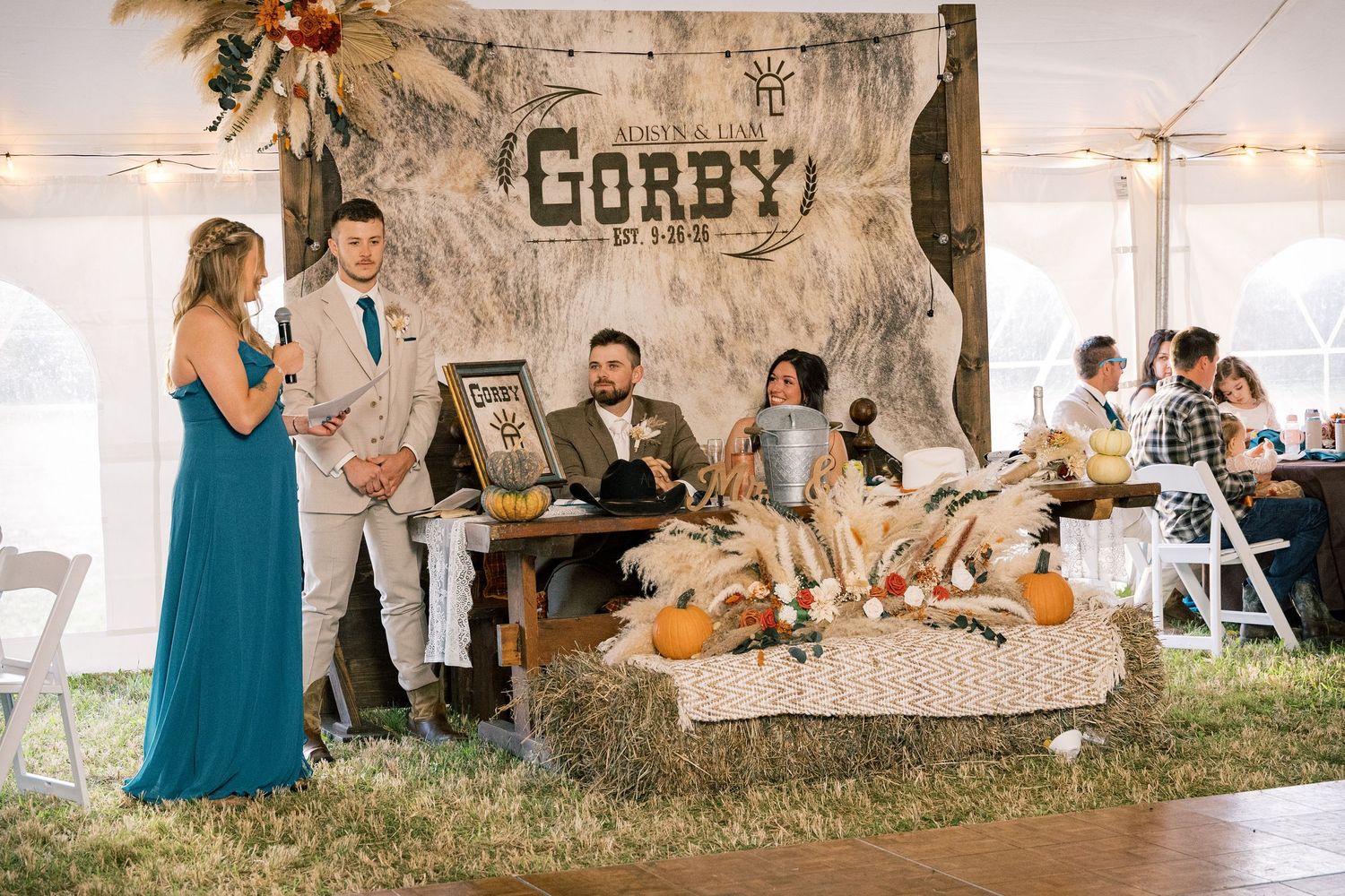 Wedding guests gather around a rustic fall-themed table with pumpkins, hay bales and a Corby sign backdrop inside a white reception tent.