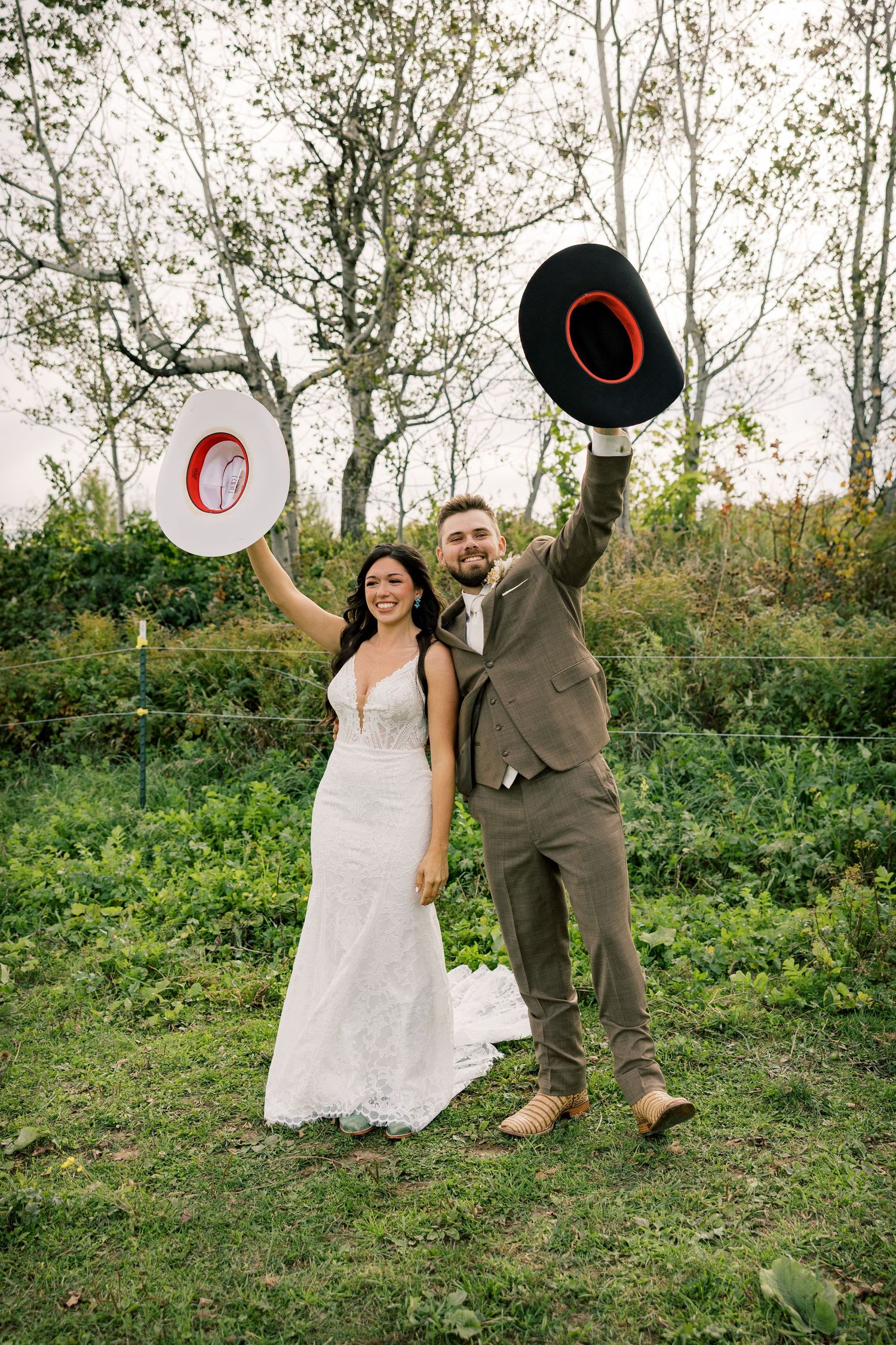 Smiling bride in a lace wedding dress and groom in a brown suit raise white and black cowboy hats in a grassy field with trees.
