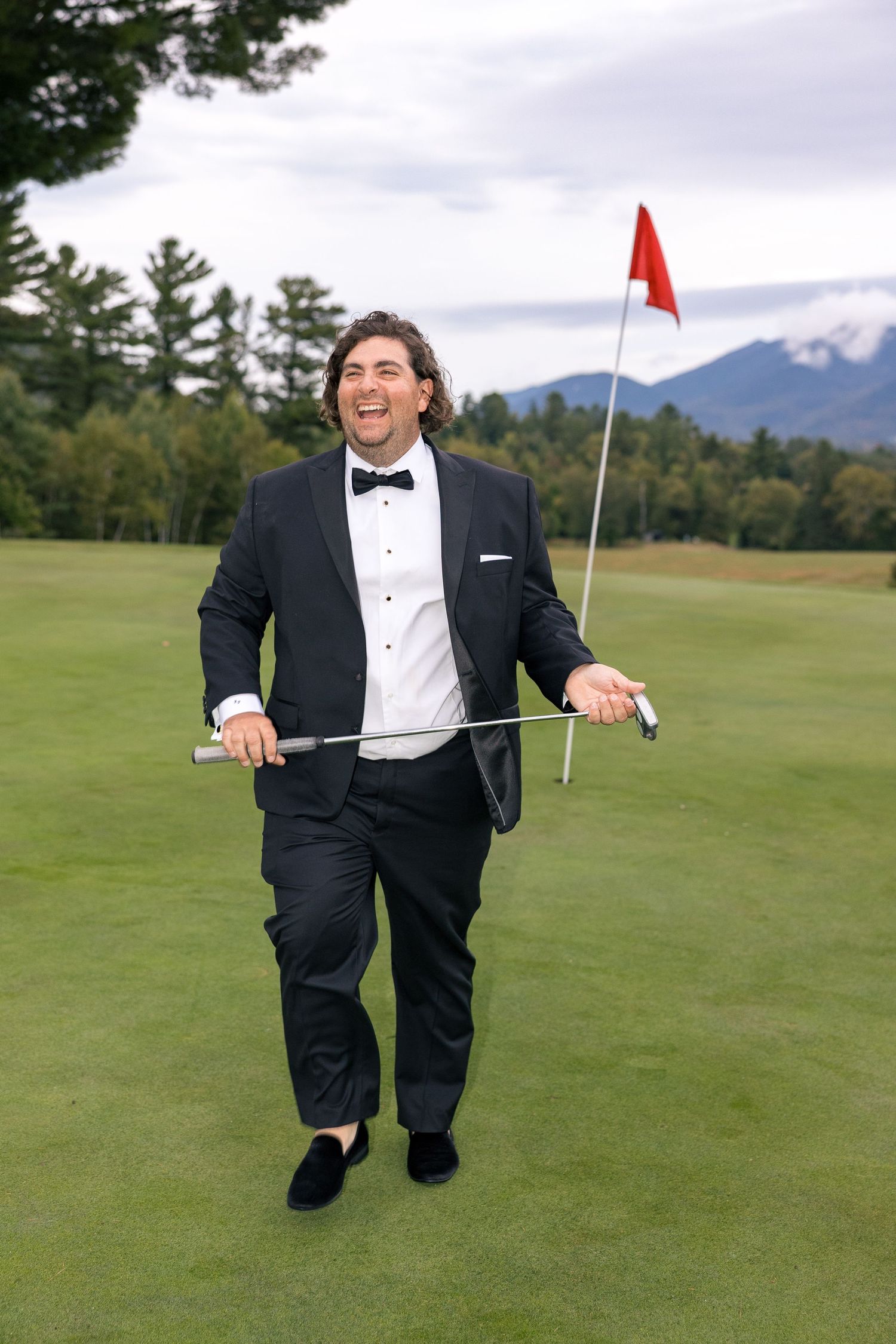 Man in black tuxedo holding golf club on green course with red flag and mountain backdrop.